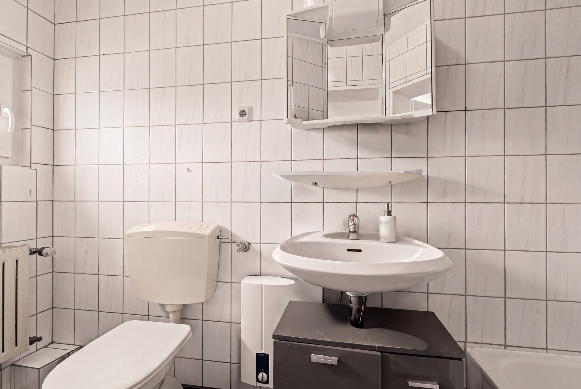 Kleines Badezimmer mit WC, Waschbecken, Spiegel und weißen Fliesen an der Wand.