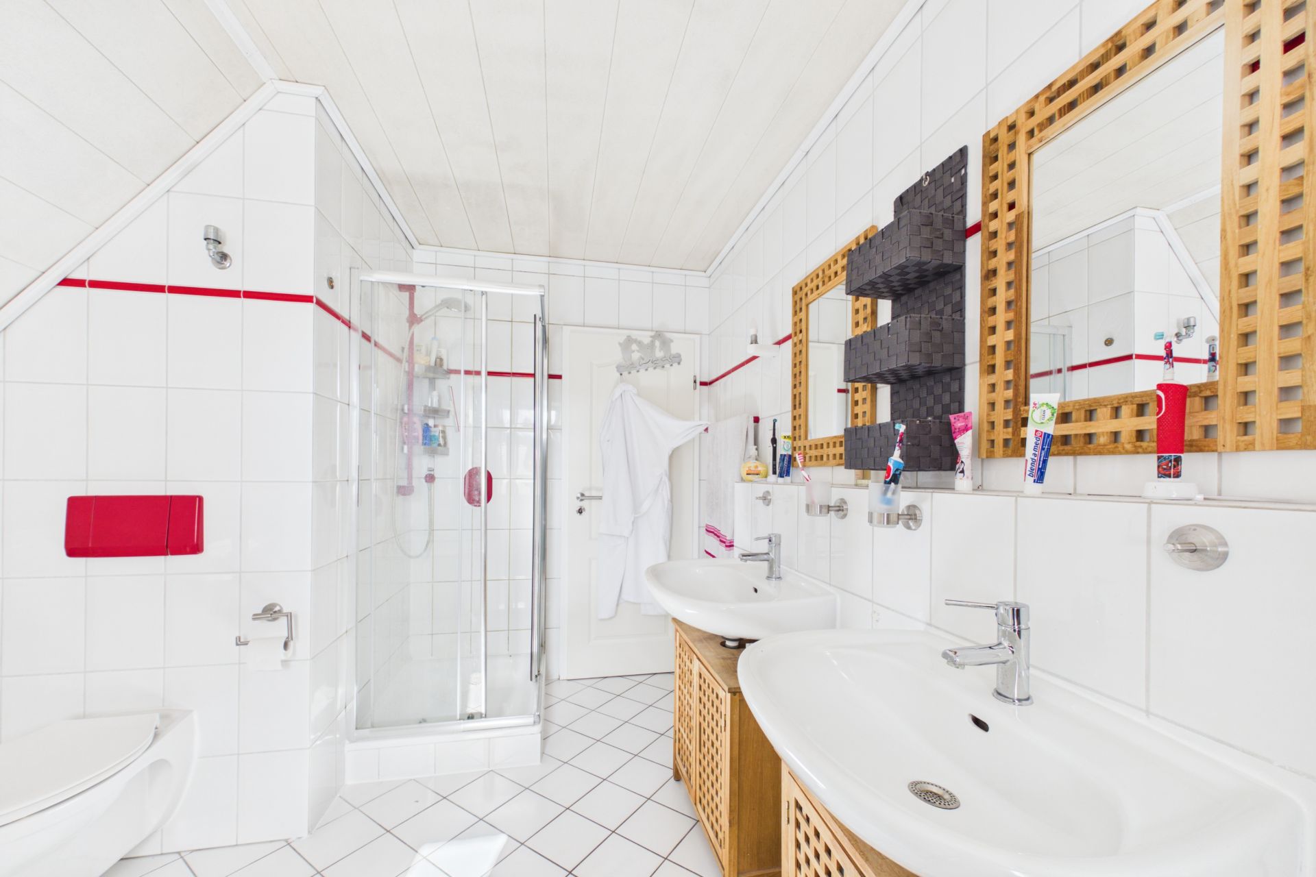 Helles Badezimmer mit Dusche, zwei Waschbecken, Spiegeln und weißen Fliesen mit rotem Streifen.