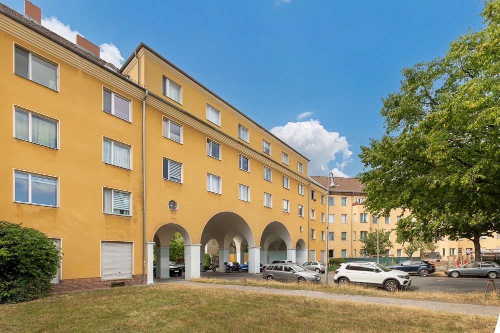 Gelbes Mehrfamilienhaus mit Rundbögen und geparkten Autos davor, daneben ein Baum und Grünfläche.