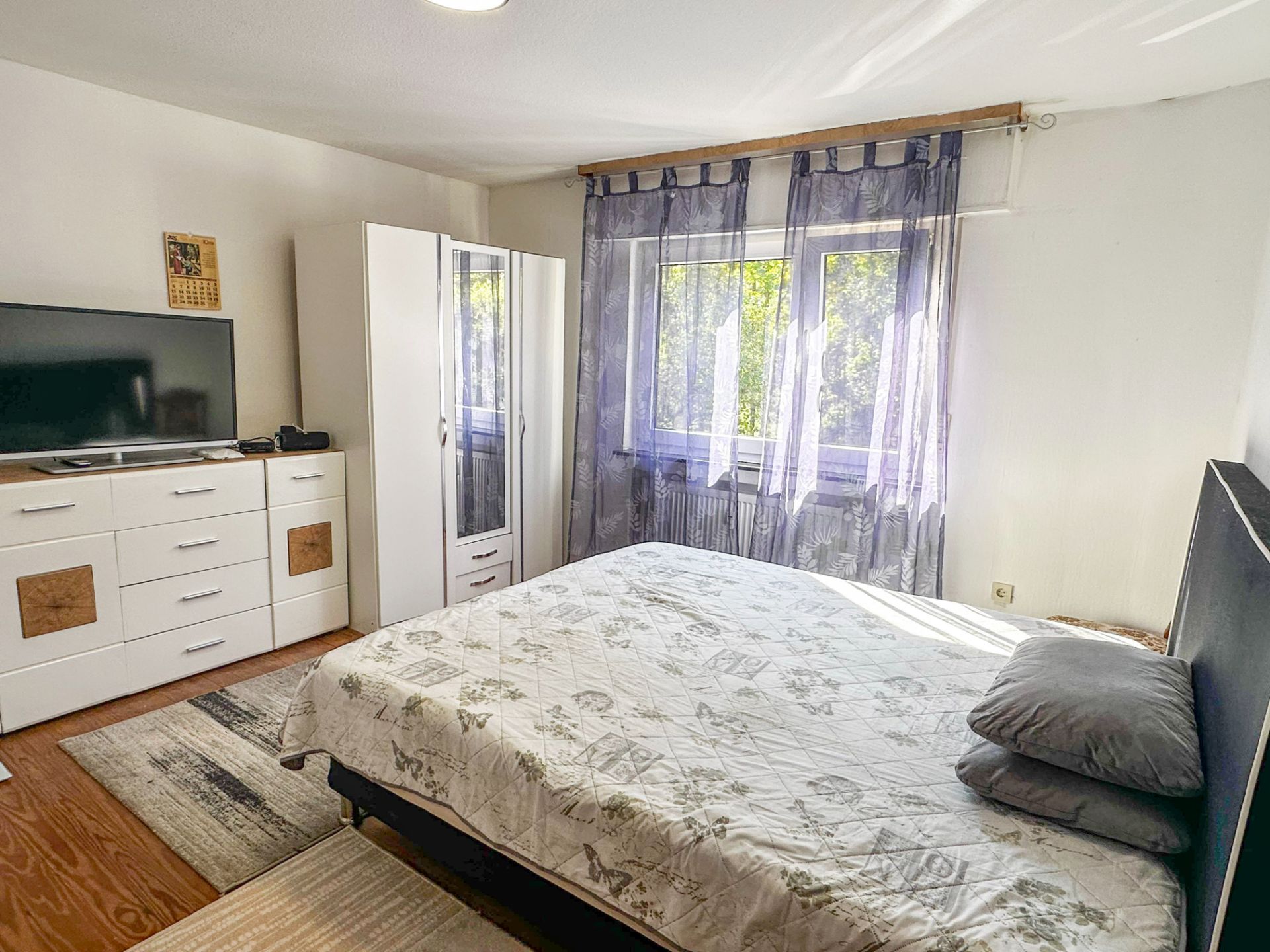 Helles Schlafzimmer mit Doppelbett, Kleiderschrank, Kommode und Fernseher am Fenster.