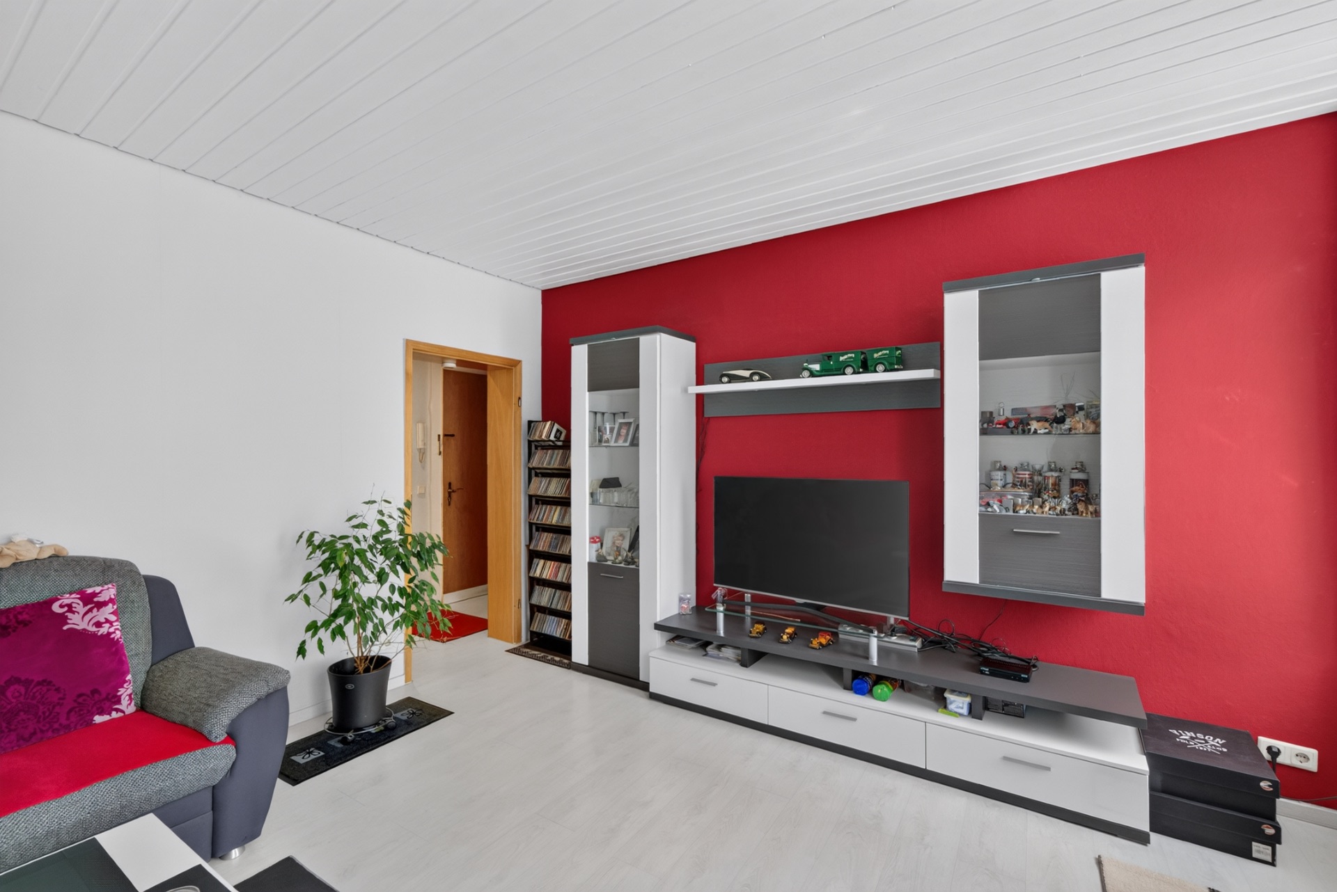 Wohnzimmer mit roter Wand, TV, Regalwand und grauem Sofa mit roten Kissen.
