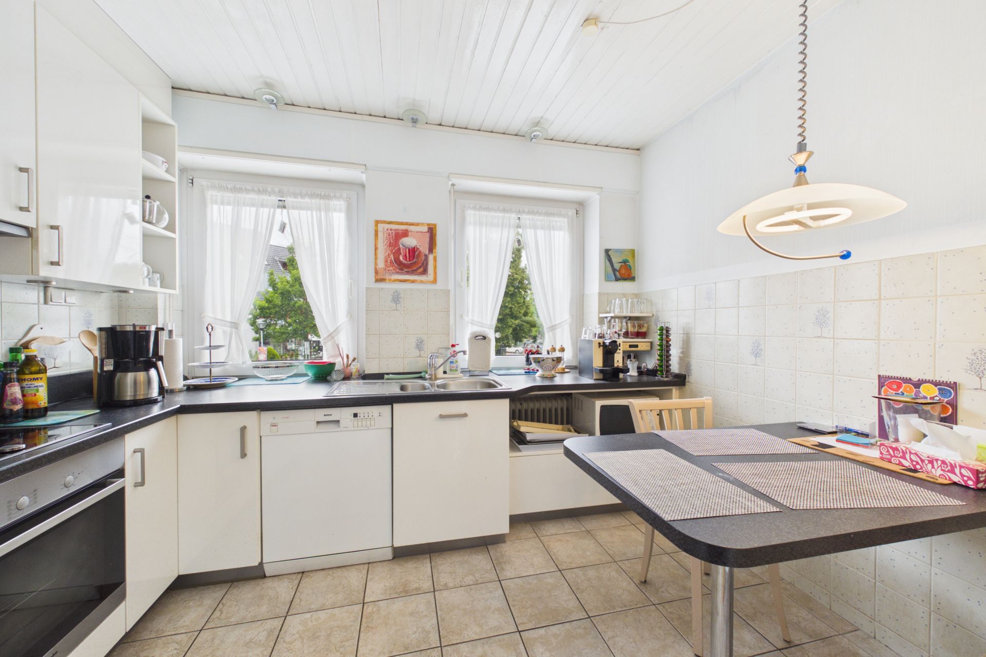Helle Küche mit weißen Schränken, Esstisch und zwei Fenstern mit Blick nach draußen.