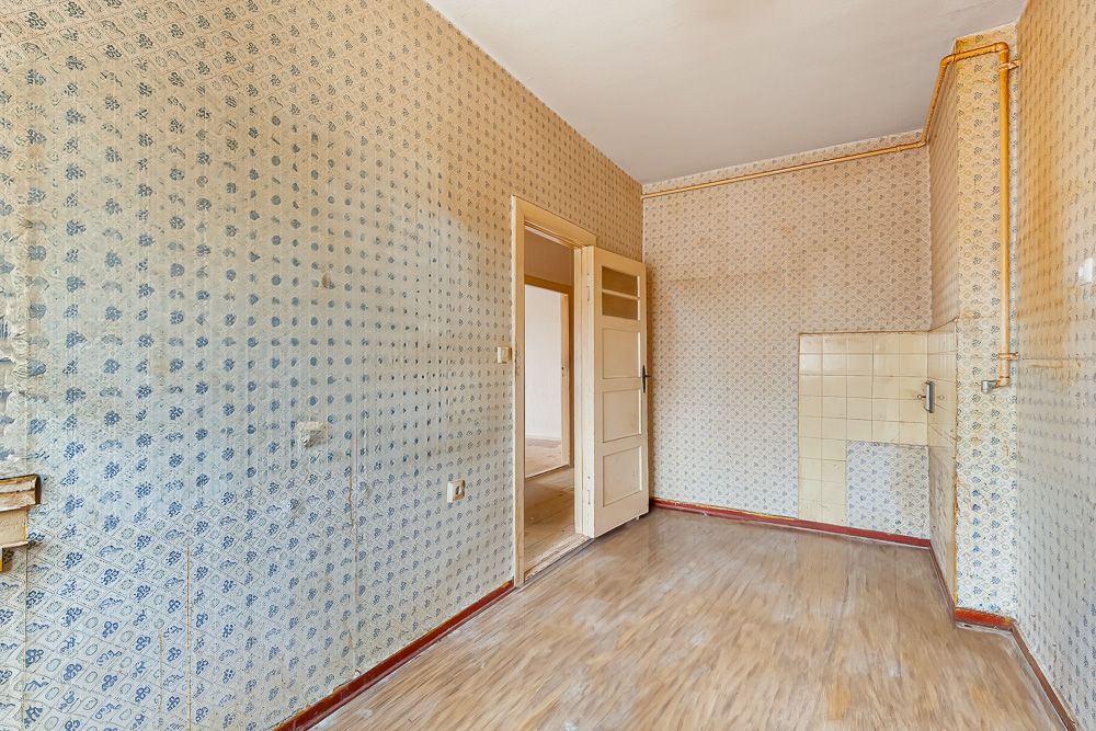 Leerer Raum mit gemusterter Tapete, Holzboden und offener Tür zu einem weiteren Zimmer.