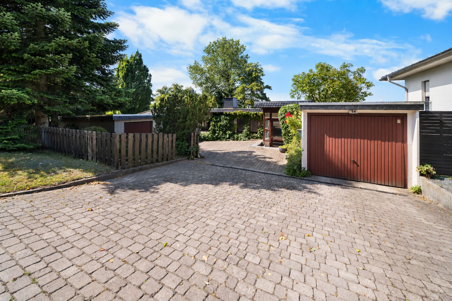 Gepflasterte Einfahrt mit Garage, Zaun, Bäumen und einem Haus im Hintergrund.