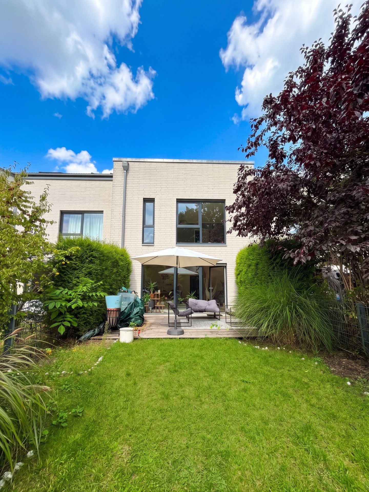 Moderner Wohnhaus-Garten mit Terrasse, Sitzgruppe und Sonnenschirm, umgeben von Pflanzen.