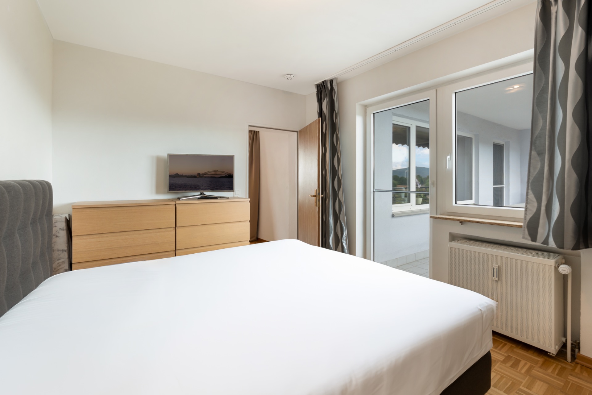 Helles Schlafzimmer mit Doppelbett, Kommode, Fernseher und Zugang zu einem Balkon.