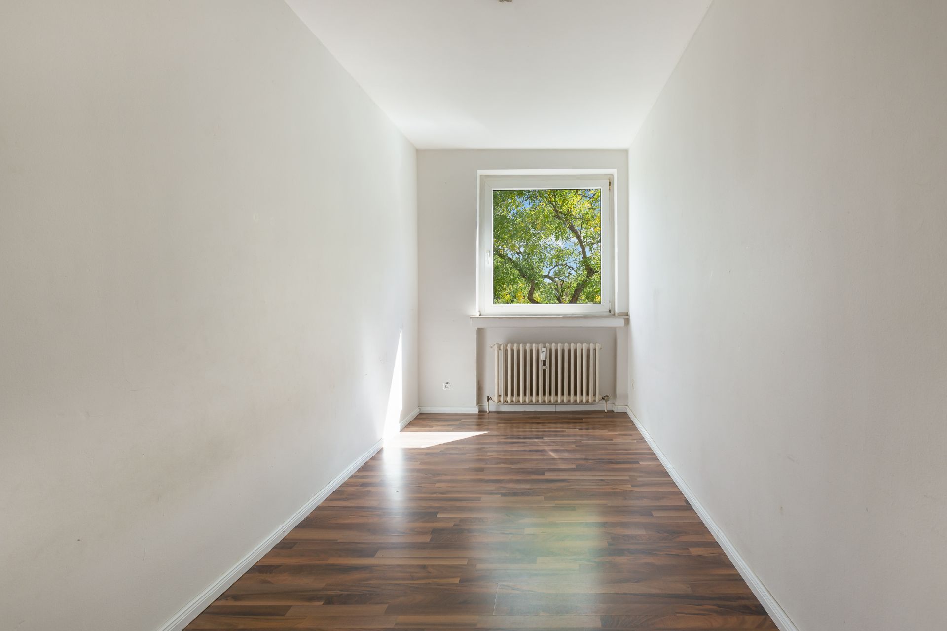 Schmaler, leerer Raum mit Holzfußboden, weißem Heizkörper und Fenster mit Blick ins Grüne.