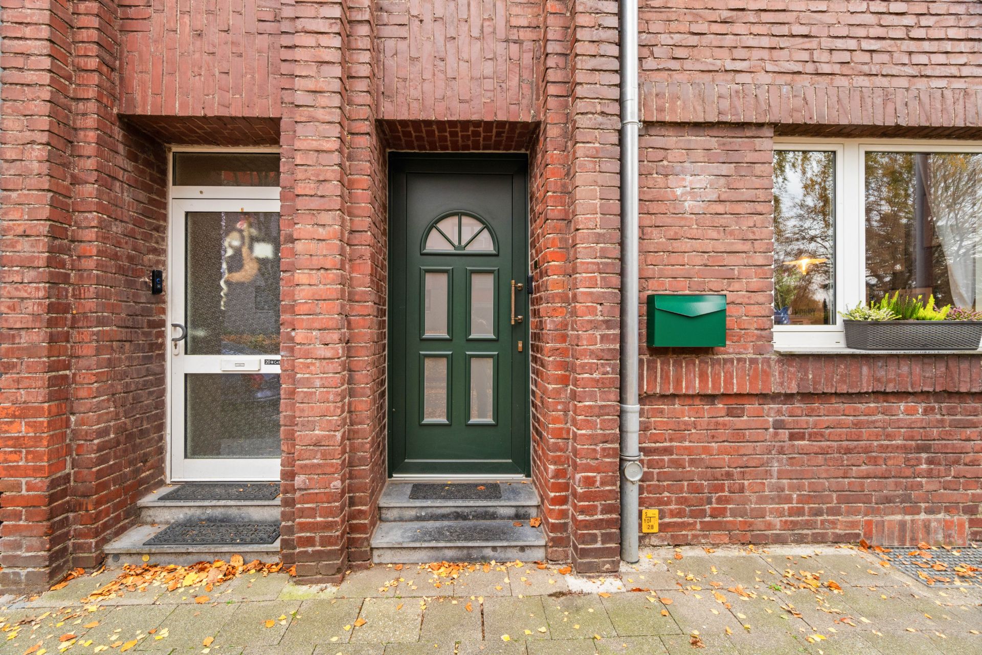 Zwei Hauseingänge mit unterschiedlichen Türen, daneben ein Fenster und ein grüner Briefkasten an einer Backsteinfassade.