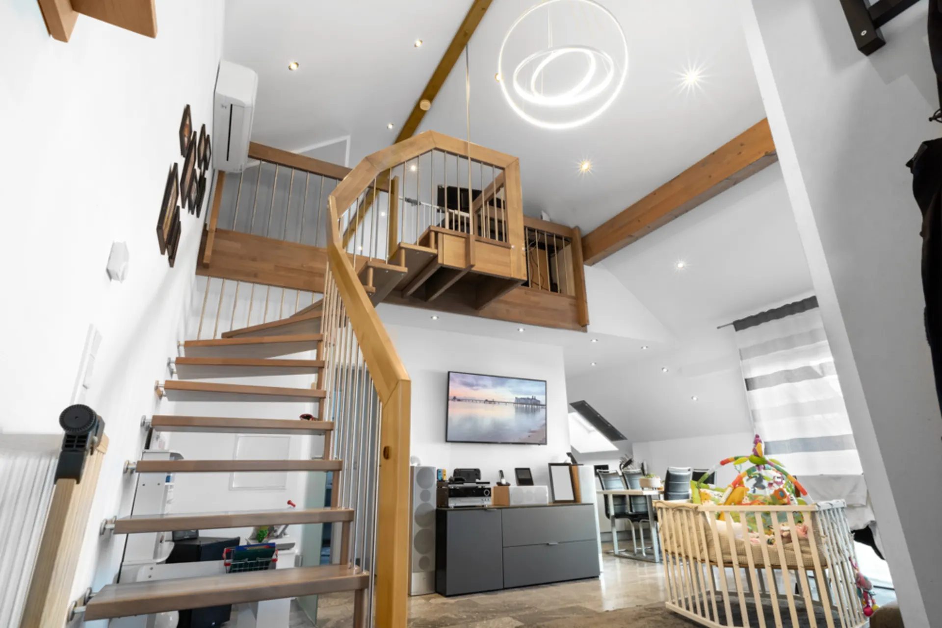 Modernes Wohnzimmer mit Holztreppe, Baby-Laufstall und Wandbild unter hoher Decke.