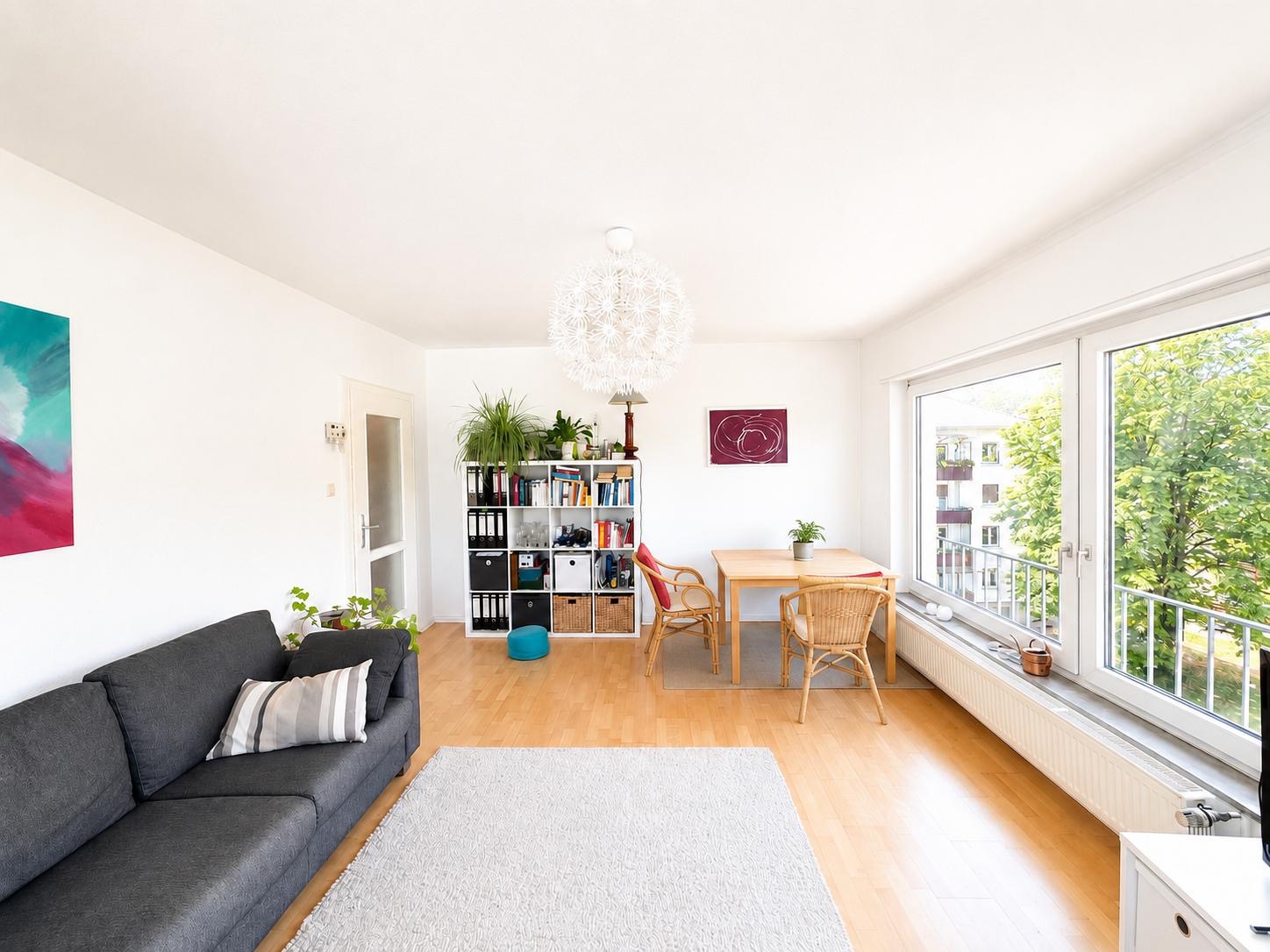 Helles Wohnzimmer mit grauem Sofa, Regal, Esstisch und großen Fenstern mit Blick ins Grüne.