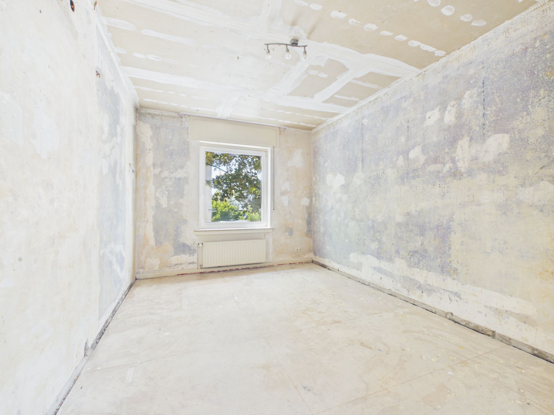 Leerer, renovierungsbedürftiger Raum mit Fenster und Heizkörper, kahle Wände und Decke.