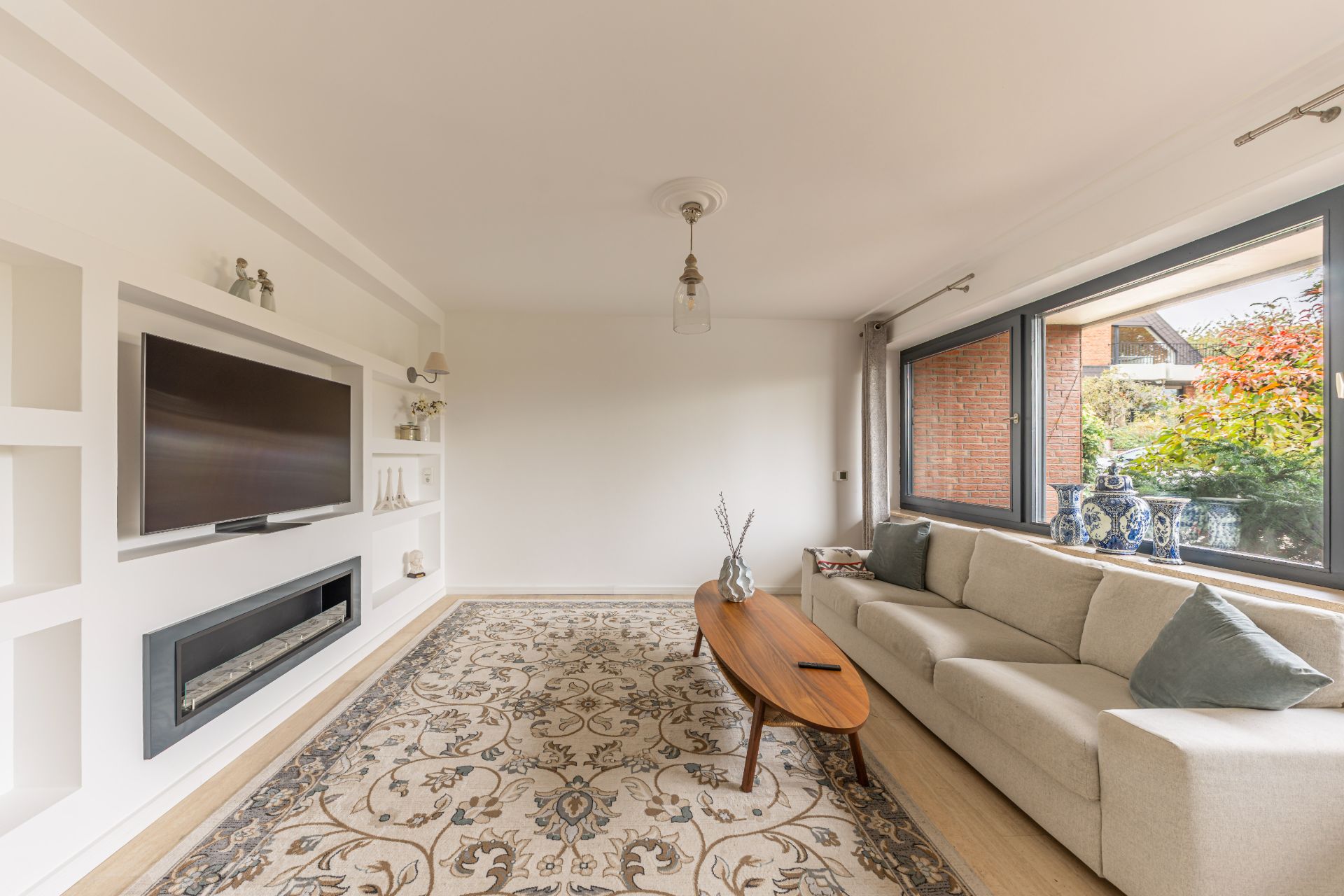 Helles Wohnzimmer mit Sofa, Teppich, Couchtisch, großem Fenster und Fernseher an der Wand.