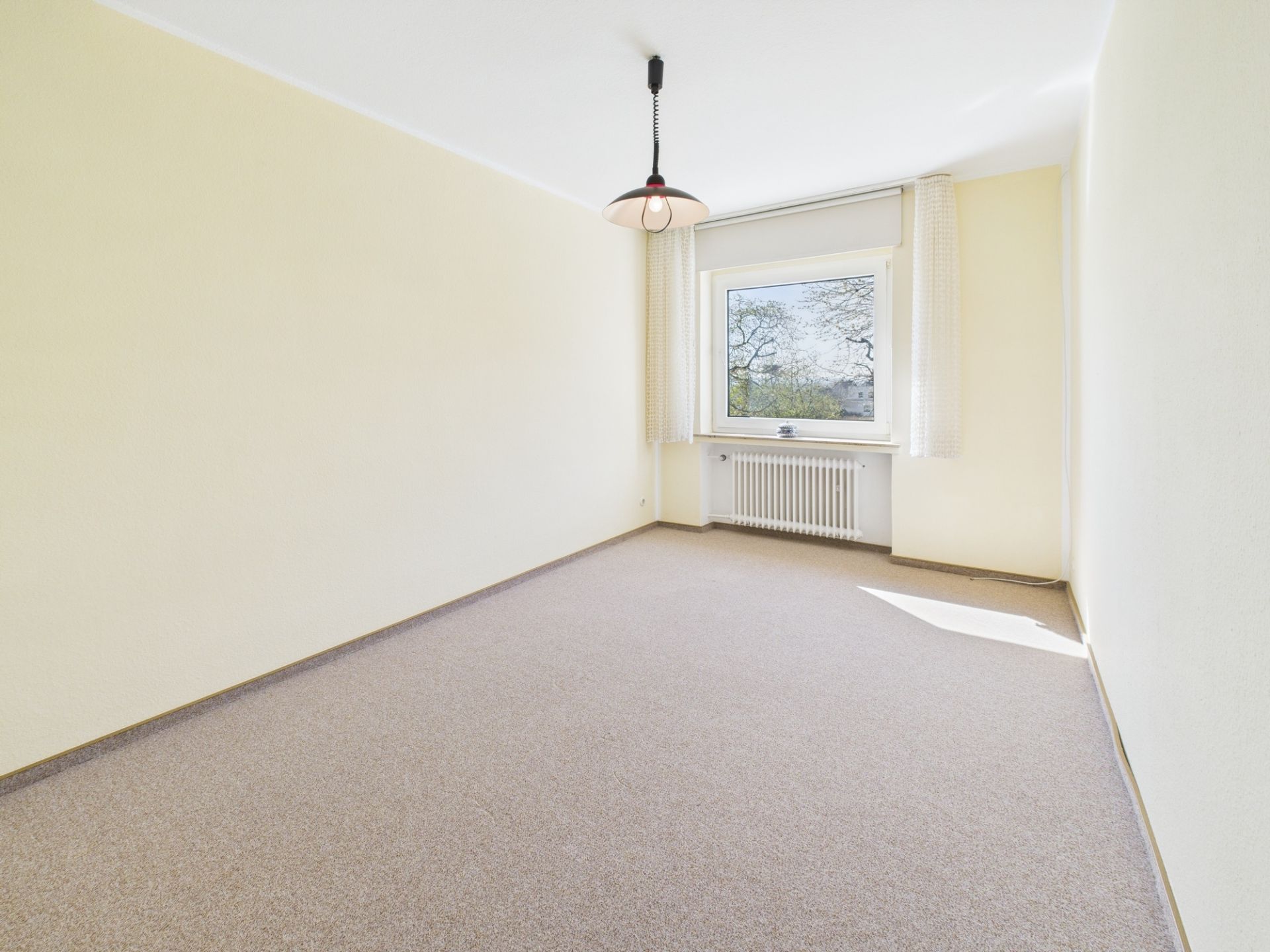 Leeres Zimmer mit Teppichboden, großem Fenster, Vorhängen und Deckenlampe.
