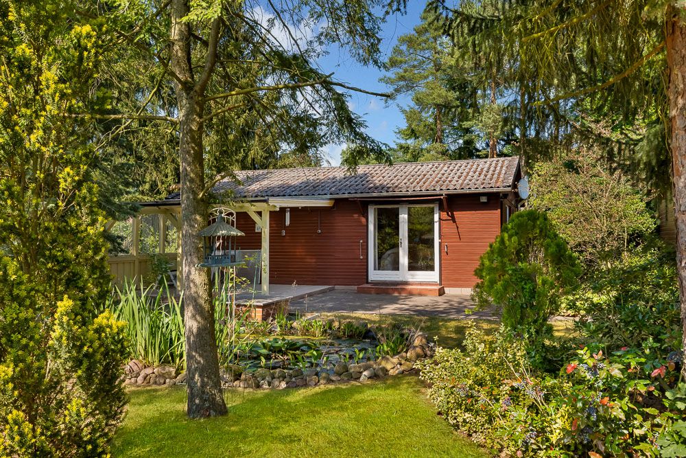 Kleines Holzhaus mit Terrasse, umgeben von Bäumen und Garten mit Teich.