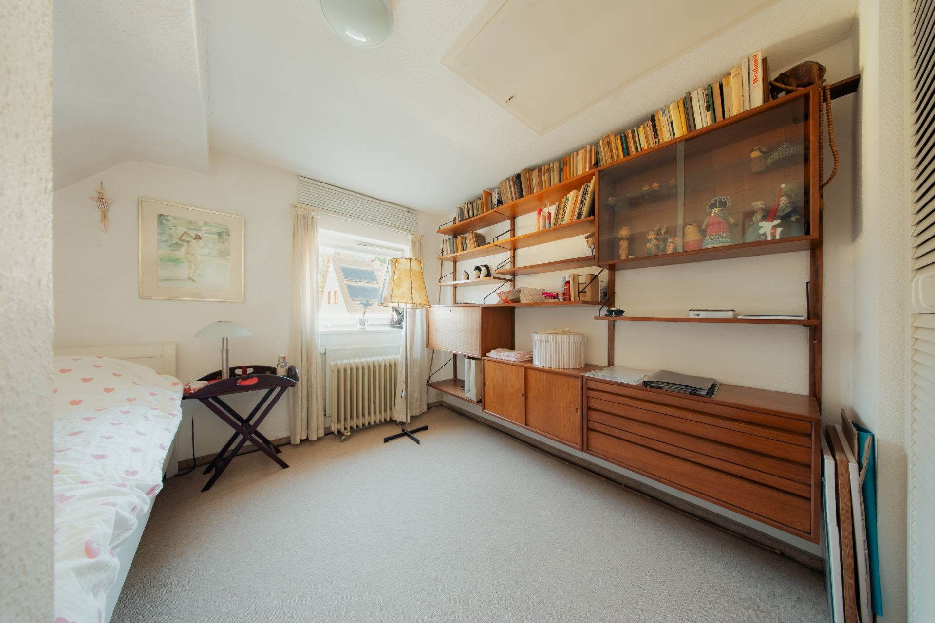 Helles Zimmer mit Einzelbett, großem Regal, Fenster und Teppichboden.