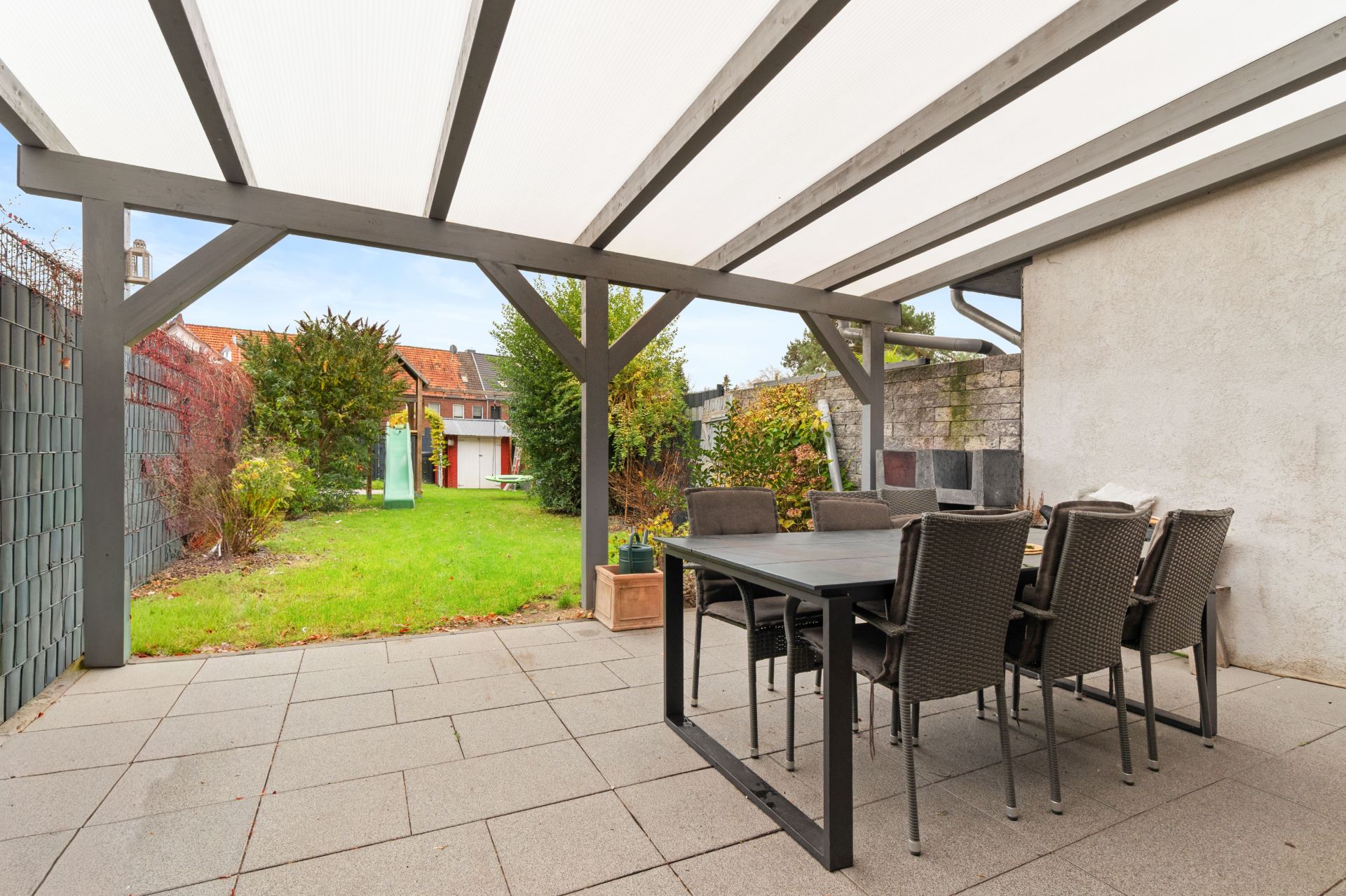 Überdachte Terrasse mit Esstisch und Stühlen, Blick auf Garten mit Rasen und Sträuchern.