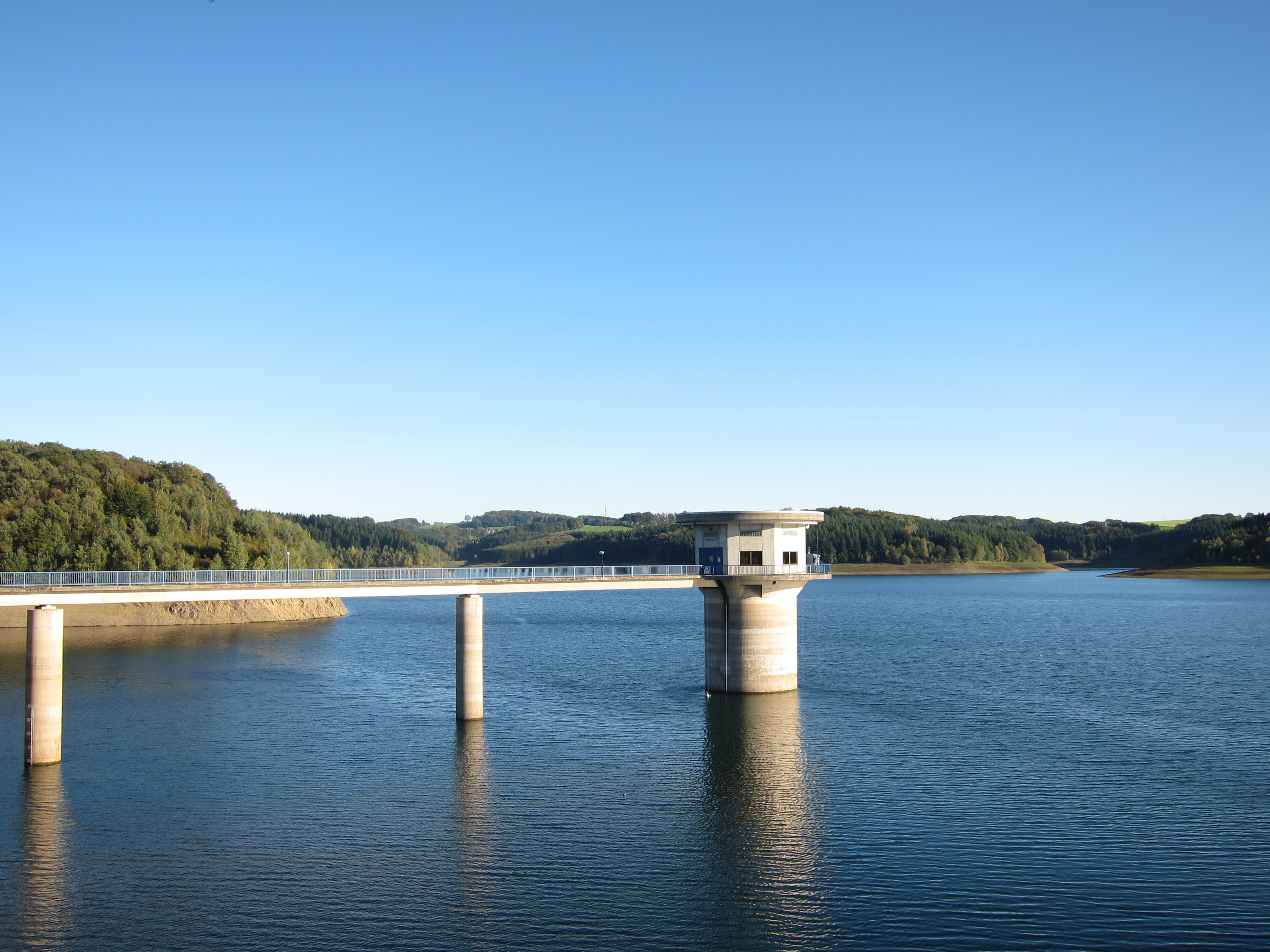 Eine Brücke mit einem kleinen Gebäude führt über einen großen See, umgeben von grünen Hügeln.