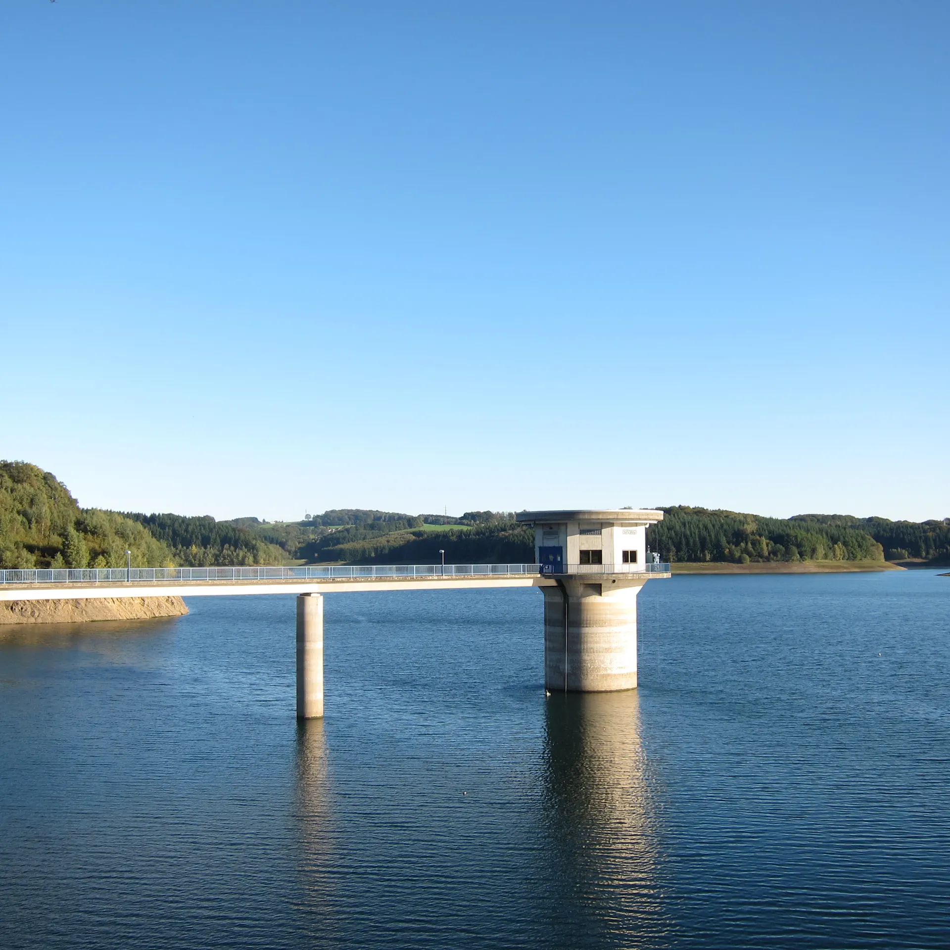 Eine Brücke mit einem kleinen Gebäude führt über einen großen See, umgeben von grünen Hügeln.