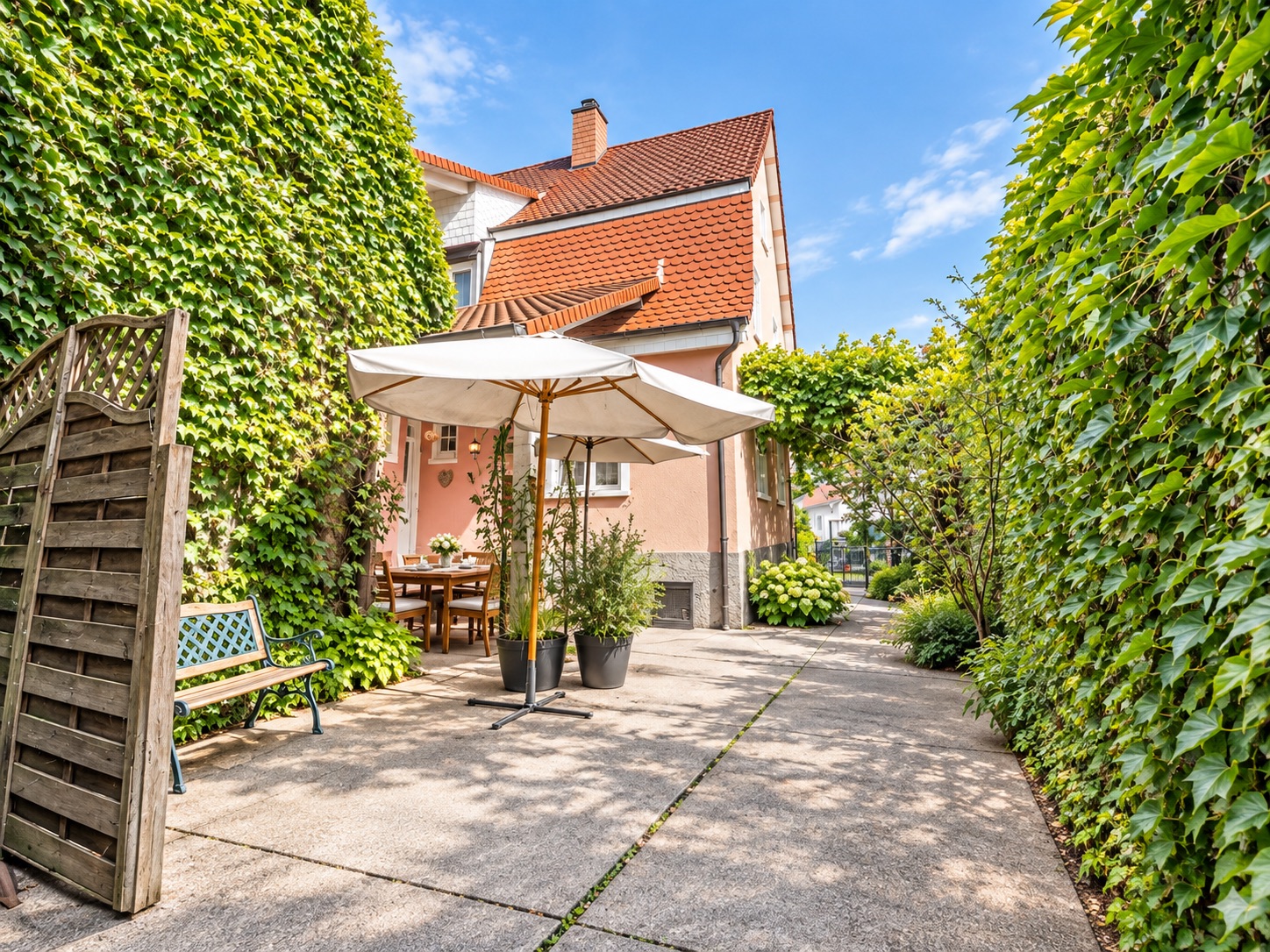 Gartenbereich mit Sonnenschirm, Bank, Esstisch und viel grünem Bewuchs an einem Wohnhaus.