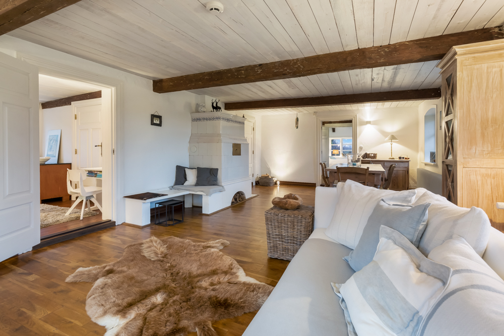 Gemütliches Wohnzimmer mit Holzbalken, weißem Sofa, Kamin und Essbereich im Hintergrund.