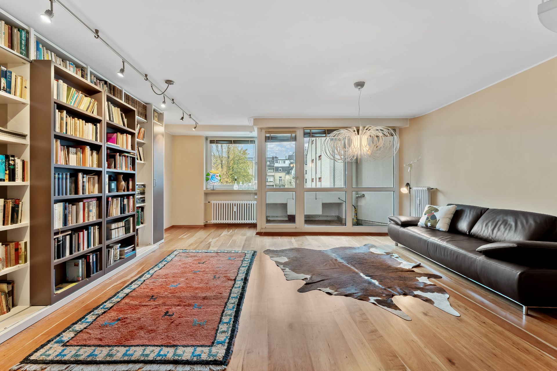 Helles Wohnzimmer mit Bücherregal, Ledersofa, Teppichen und großen Fenstern.