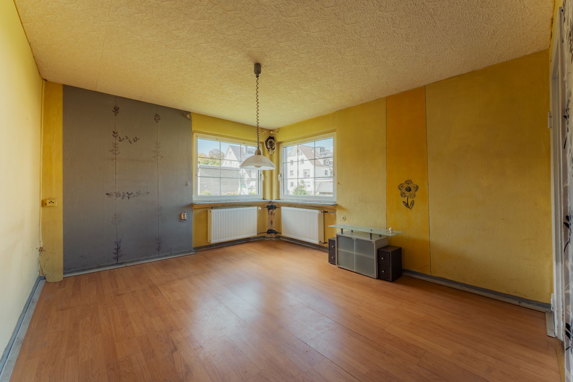 Leeres Zimmer mit gelben Wänden, zwei Fenstern, Holzfußboden und einer Deckenlampe.