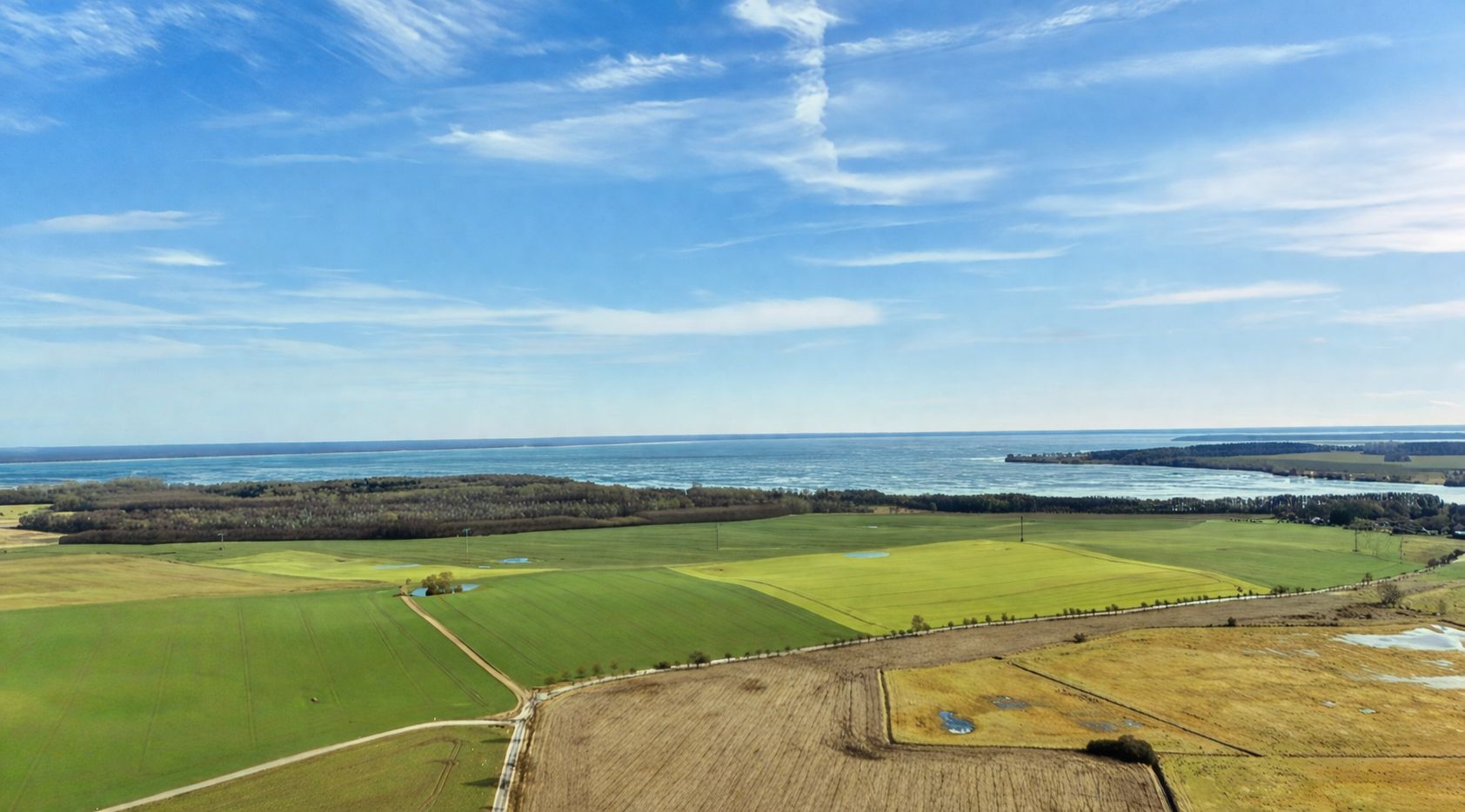 Landschaft mit Feldern, Wiesen und Blick auf ein großes Gewässer unter blauem Himmel.