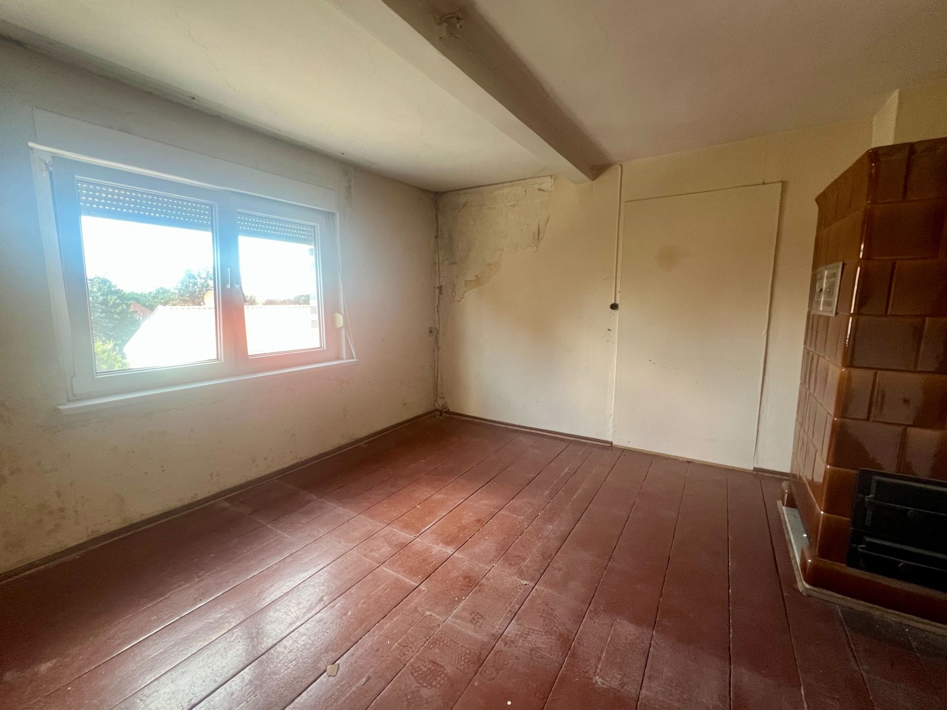 Leerer Raum mit braunem Holzboden, Fenster, Ofen und abgenutzter Wand.