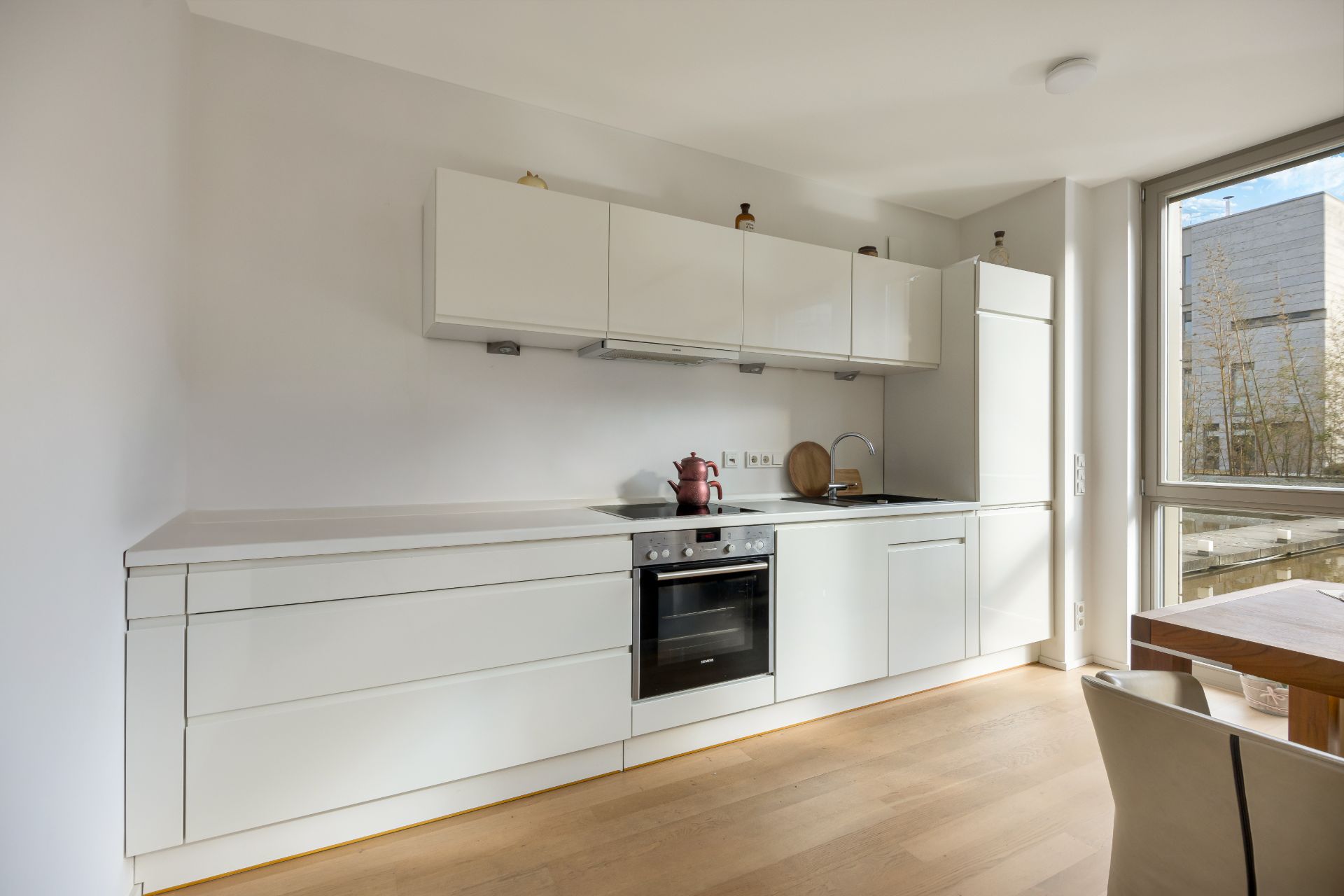 Moderne, weiße Einbauküche mit Herd, Ofen, Spüle und großem Fenster, daneben ein Esstisch.