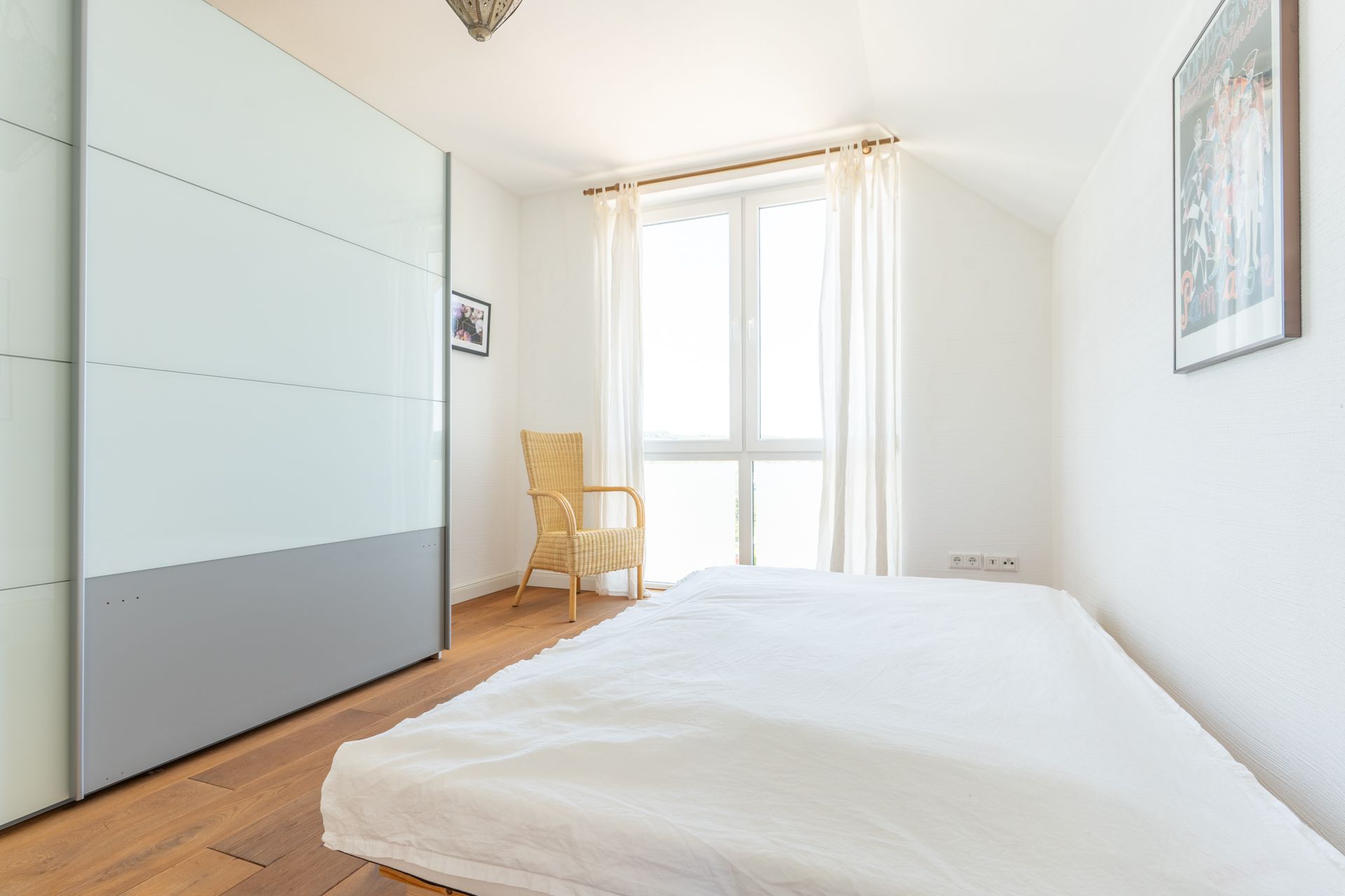 Helles Schlafzimmer mit niedrigem Bett, großem Schrank, Fenster mit Vorhängen und Stuhl.