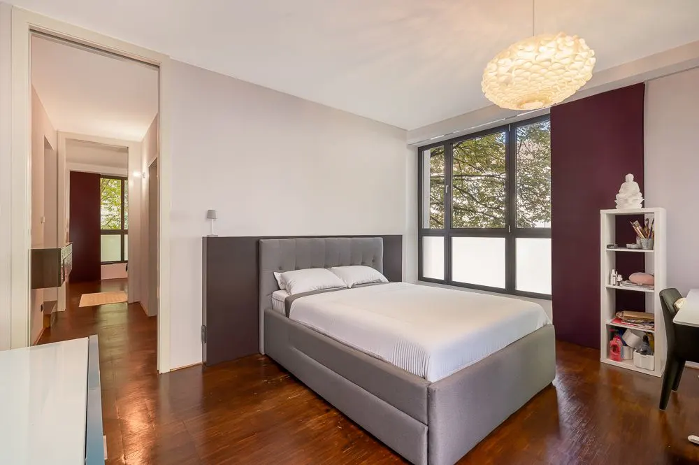 Modernes Schlafzimmer mit großem Bett, Fenster, Regal und Schreibtisch auf Holzboden.