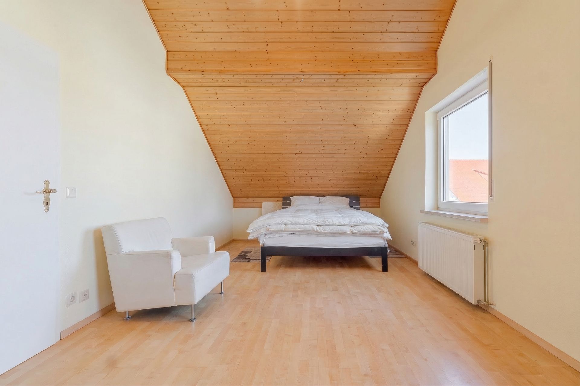 Helles Schlafzimmer mit Holzdecke, Bett, Sessel und Fenster.