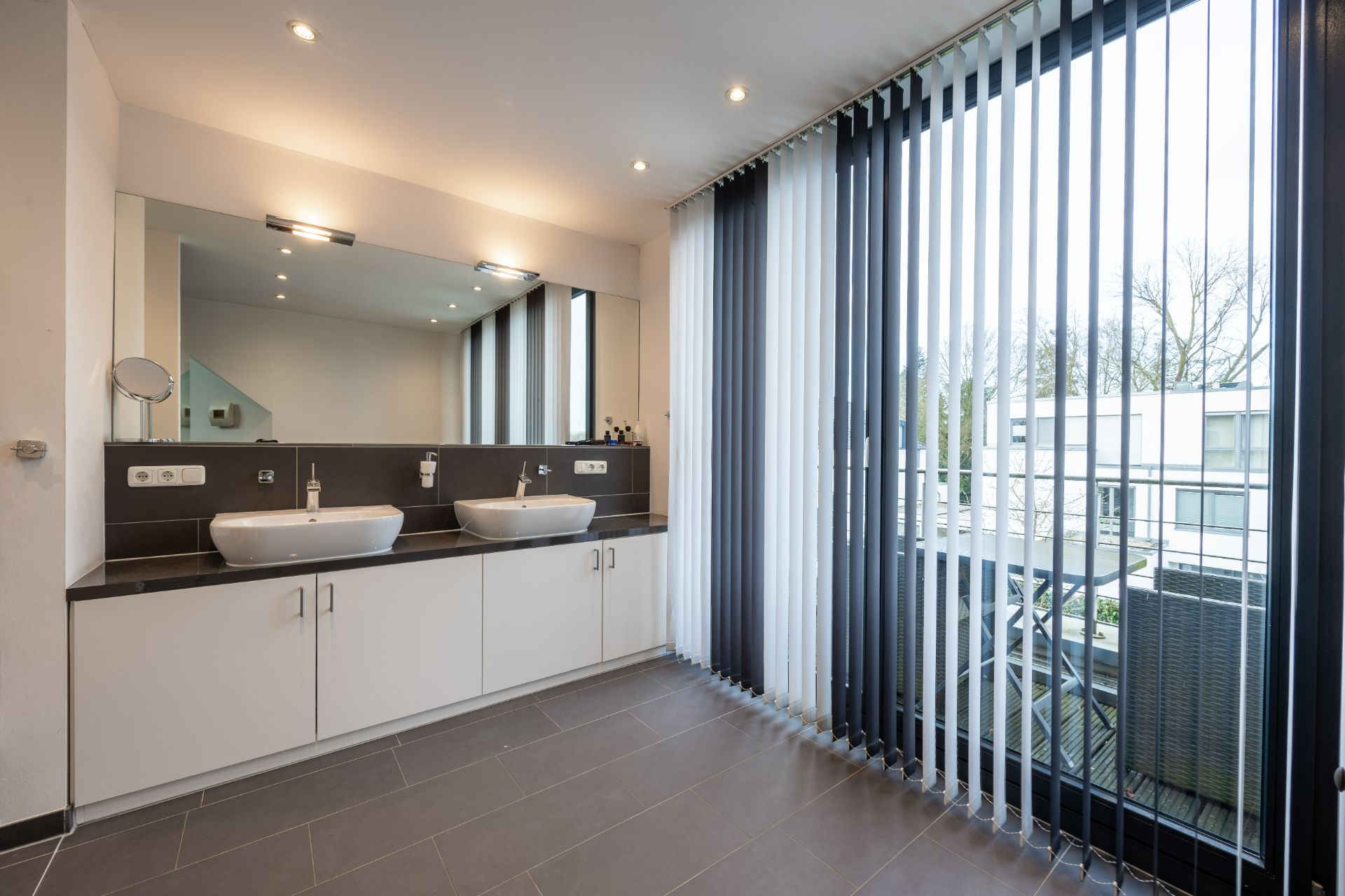 Modernes Badezimmer mit zwei Waschbecken, großem Spiegel und Fenster mit Lamellenvorhängen.