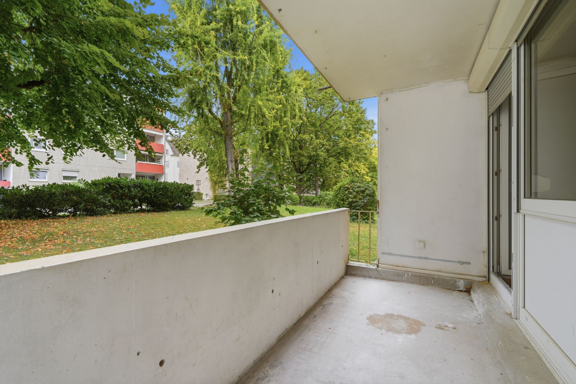 Überdachter Balkon mit Blick auf grüne Bäume und eine Rasenfläche vor einem Wohngebäude.