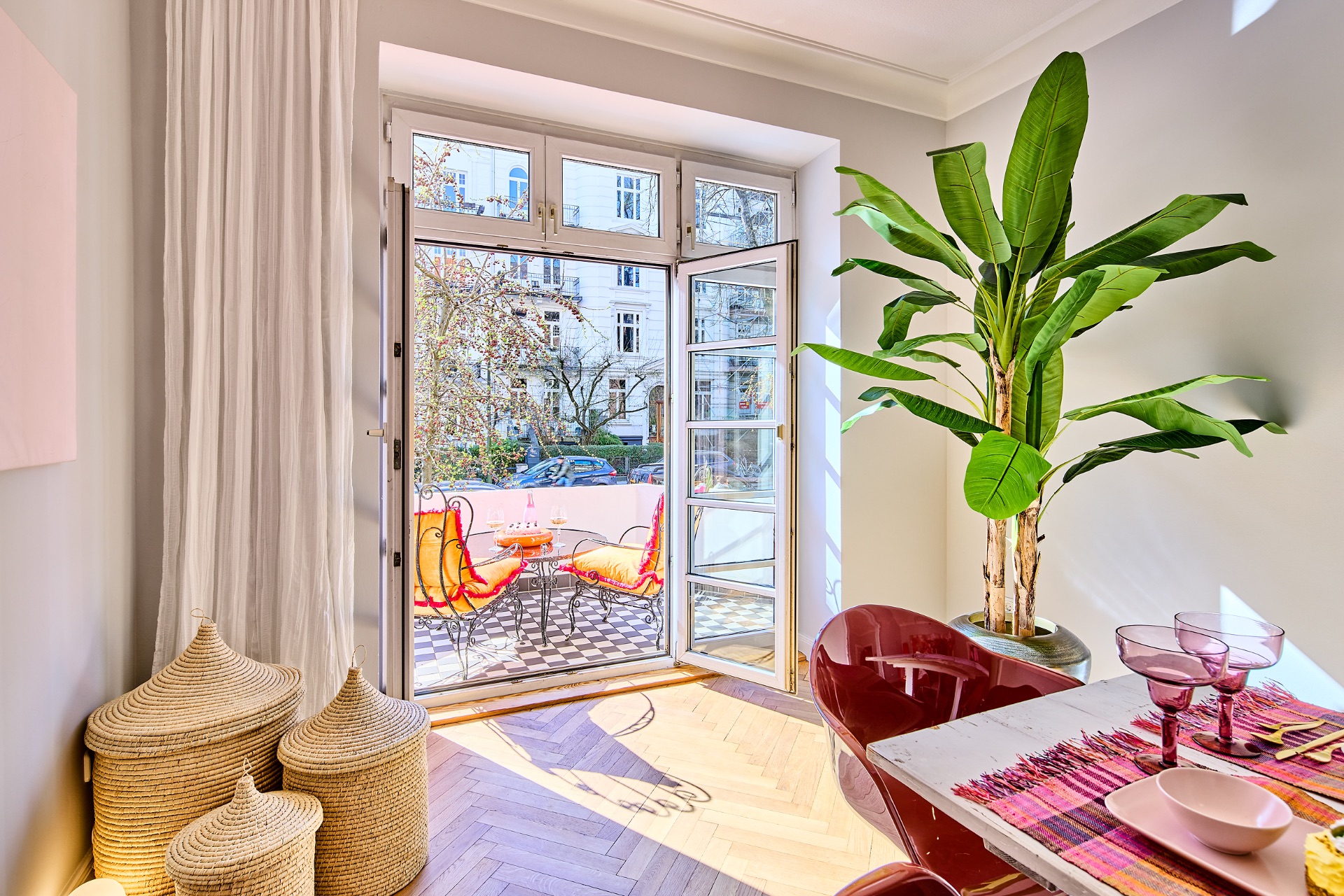 Helles Wohnzimmer mit offener Tür zum Balkon, Esstisch, großen Pflanzen und dekorativen Körben.