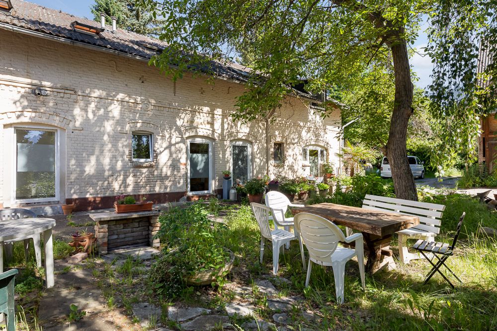 Gemütlicher Garten mit Holztisch, weißen Stühlen und Bäumen vor einem hellen Backsteinhaus.