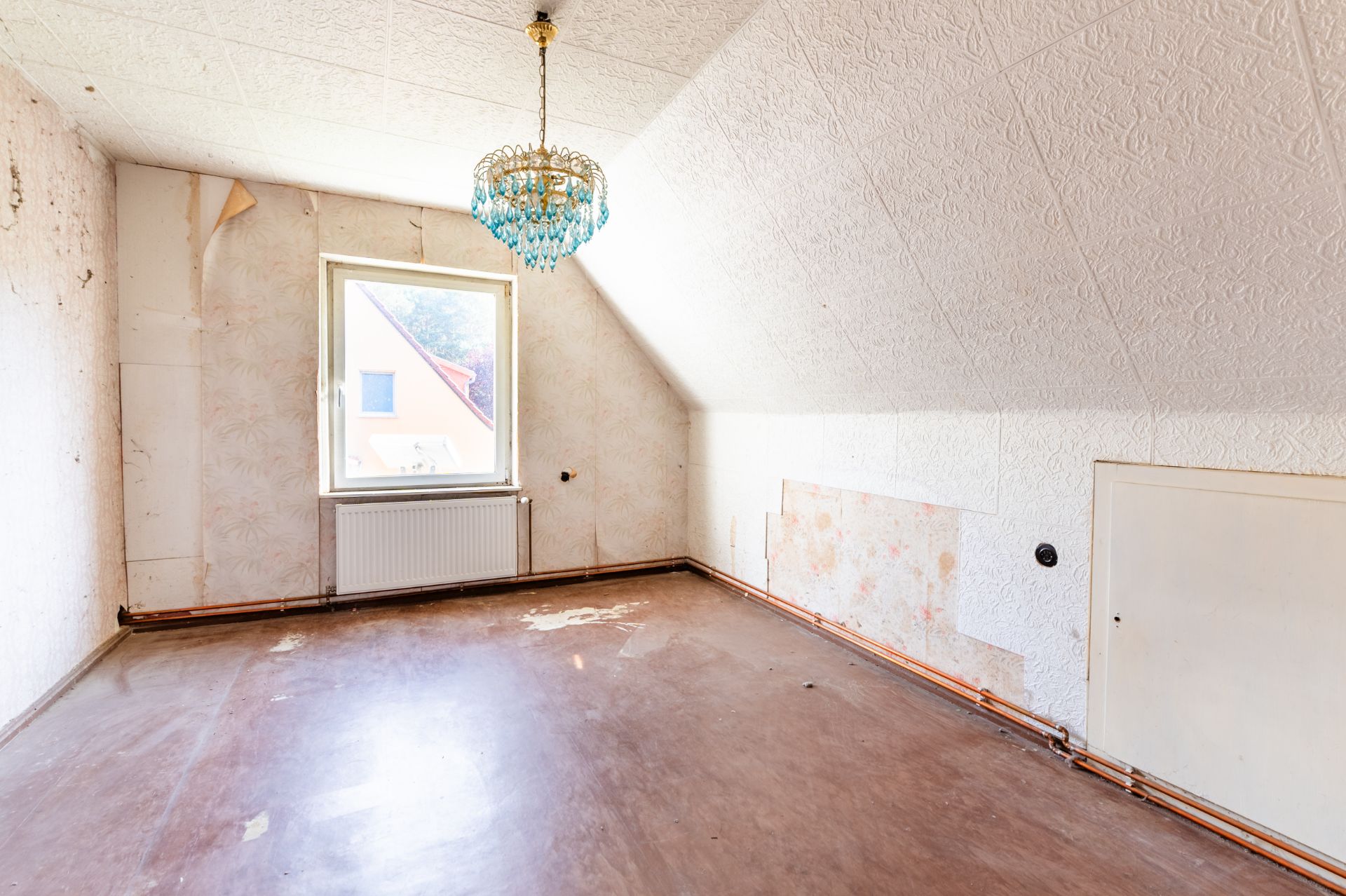 Leerer Raum mit schräger Decke, Fenster, Heizung und Kronleuchter.