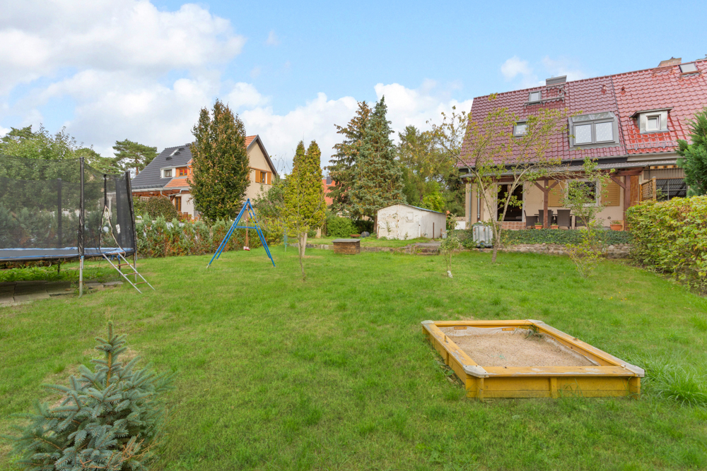 Großer Garten mit Sandkasten, Trampolin, Schaukel und Rasenfläche vor Wohnhäusern.