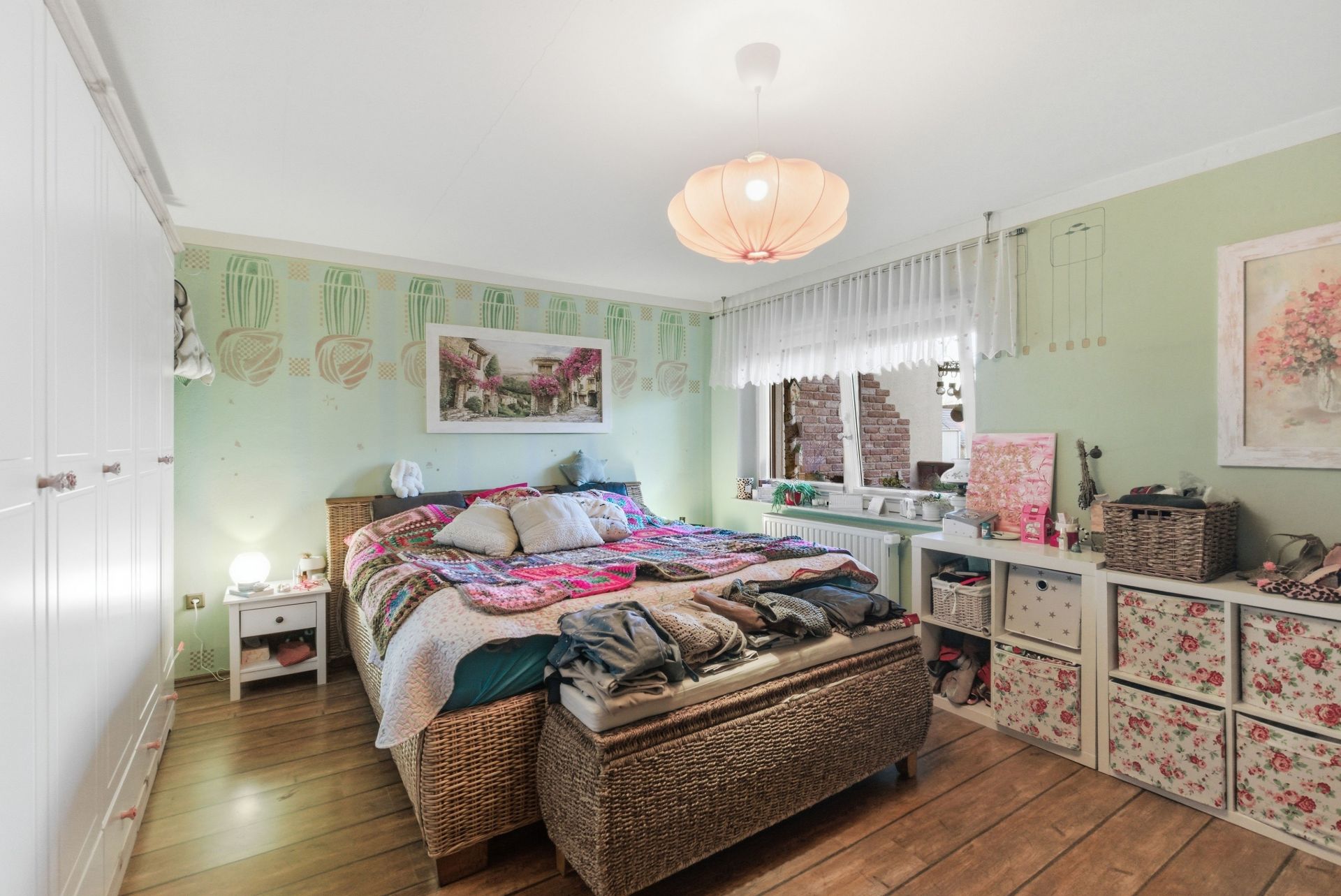 Helles Schlafzimmer mit Doppelbett, Kommode, Schrank und Fenster mit Gardinen.