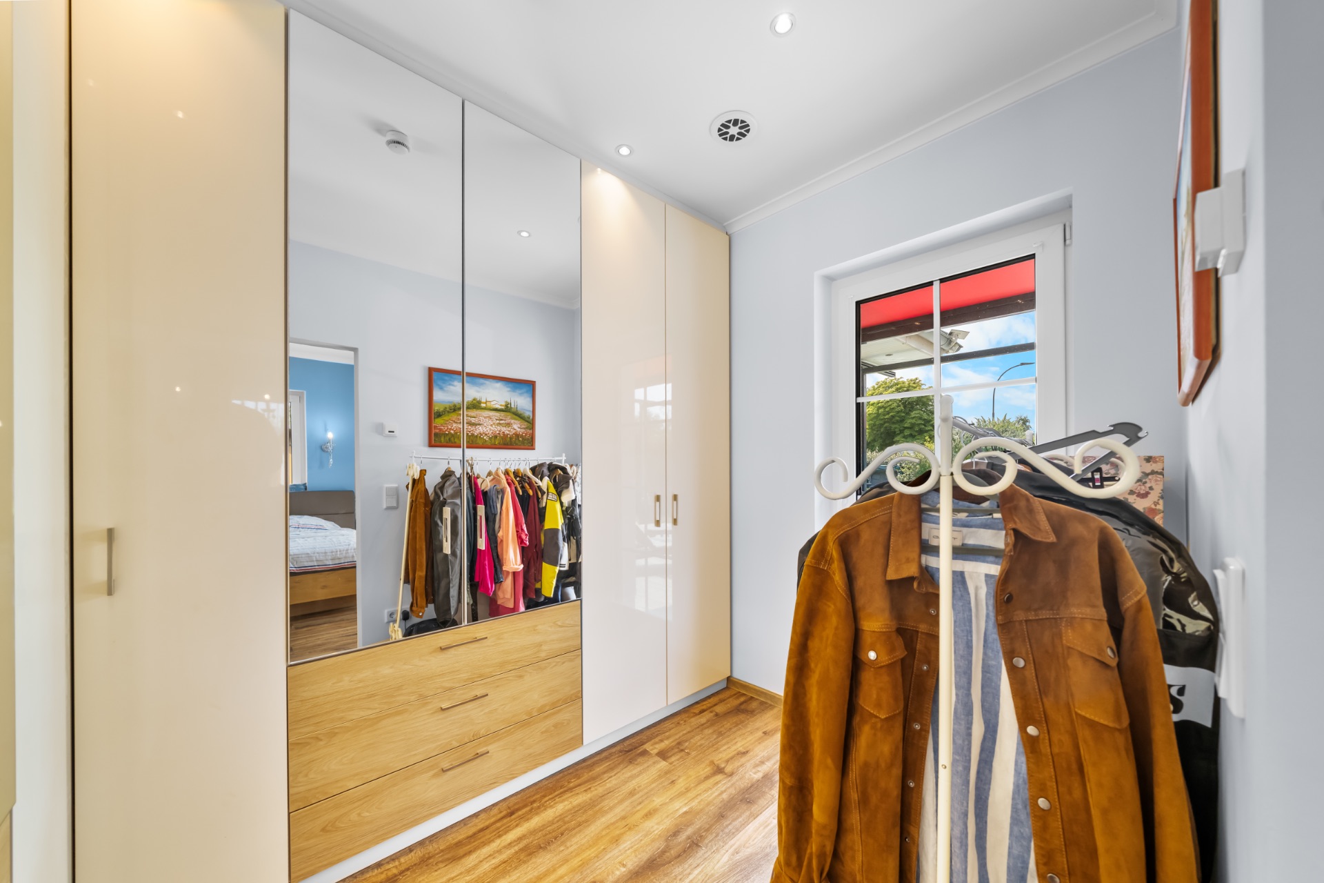 Ankleidezimmer mit Spiegel-Kleiderschrank, Fenster und Kleiderständer auf Holzfußboden.