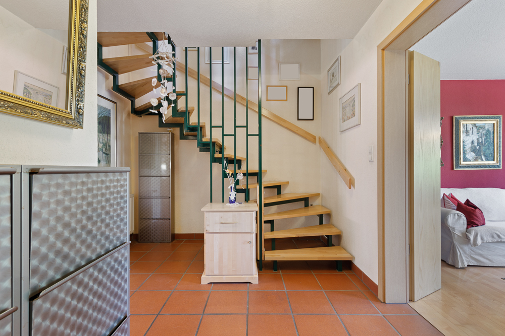Heller Flur mit Holztreppe, Fliesenboden und Zugang zum Wohnzimmer mit roter Wand.