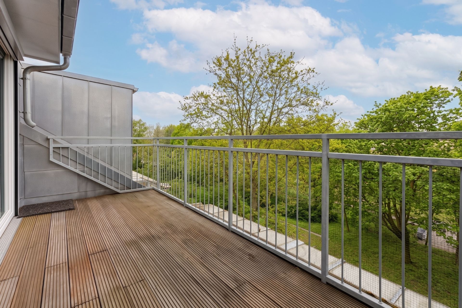 Großer Holzbalkon mit Metallgeländer, Blick auf grüne Bäume unter blauem Himmel.