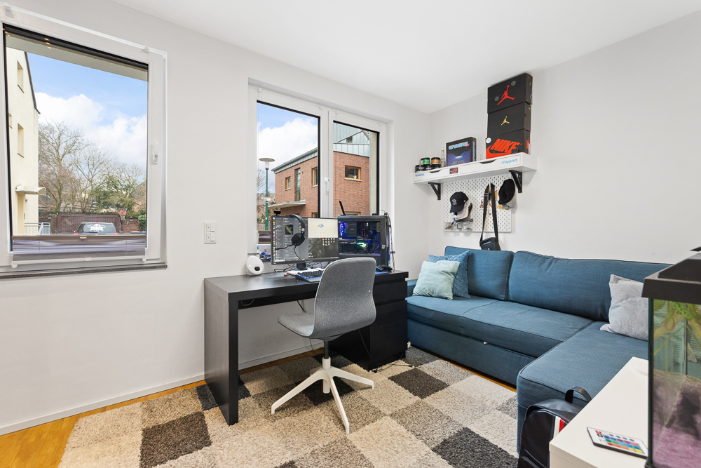 Modernes Arbeitszimmer mit Schreibtisch, Computer, grauem Stuhl und blauer Eckcouch vor zwei Fenstern.