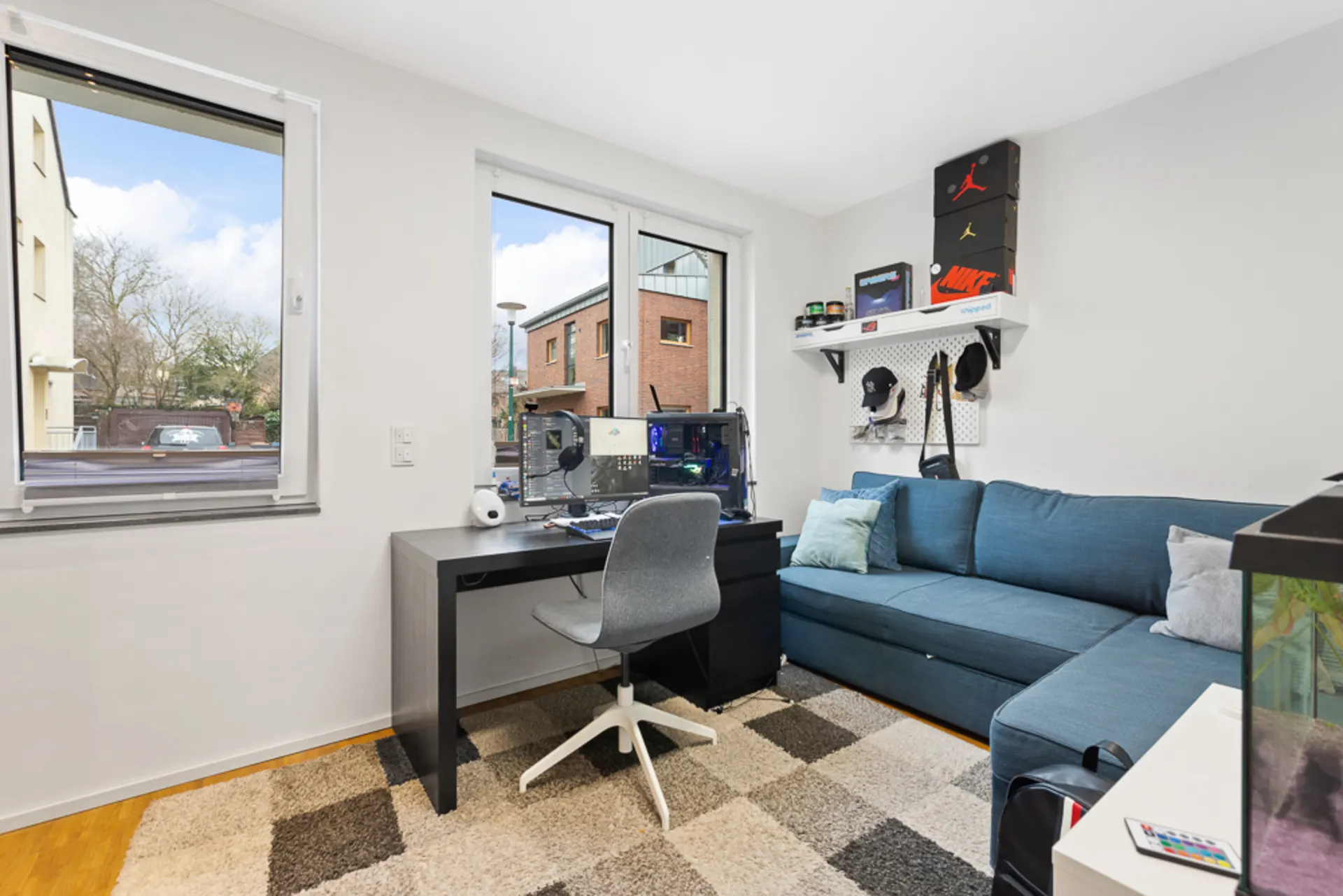 Modernes Arbeitszimmer mit Schreibtisch, Computer, grauem Stuhl und blauer Eckcouch vor zwei Fenstern.