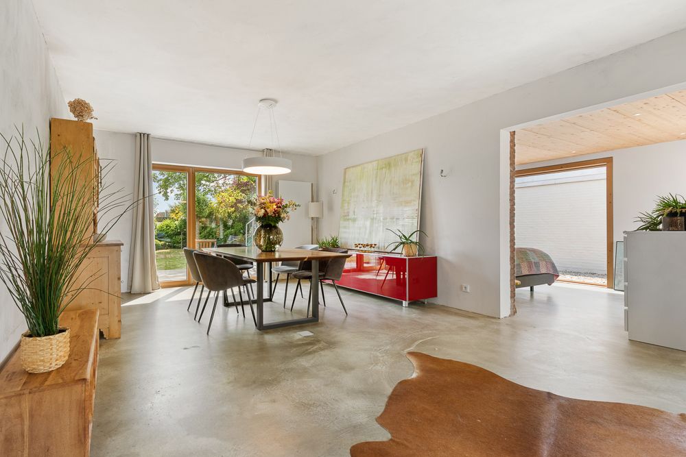 Heller Essbereich mit großem Fenster, Holzmöbeln, Pflanzen und Blick in ein Schlafzimmer.
