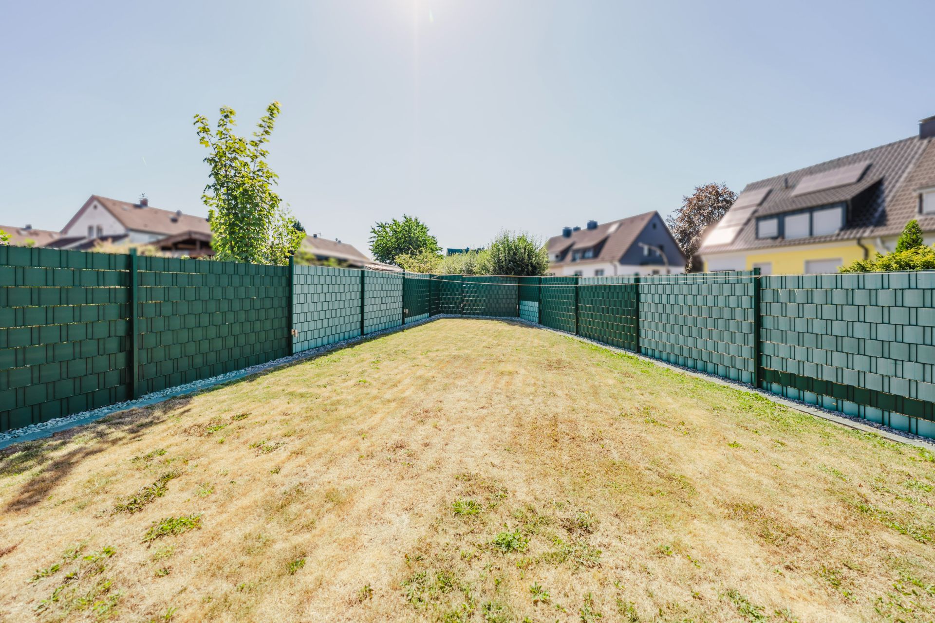 Garten mit grünem Zaun und Rasen, im Hintergrund Nachbarhäuser bei sonnigem Wetter.