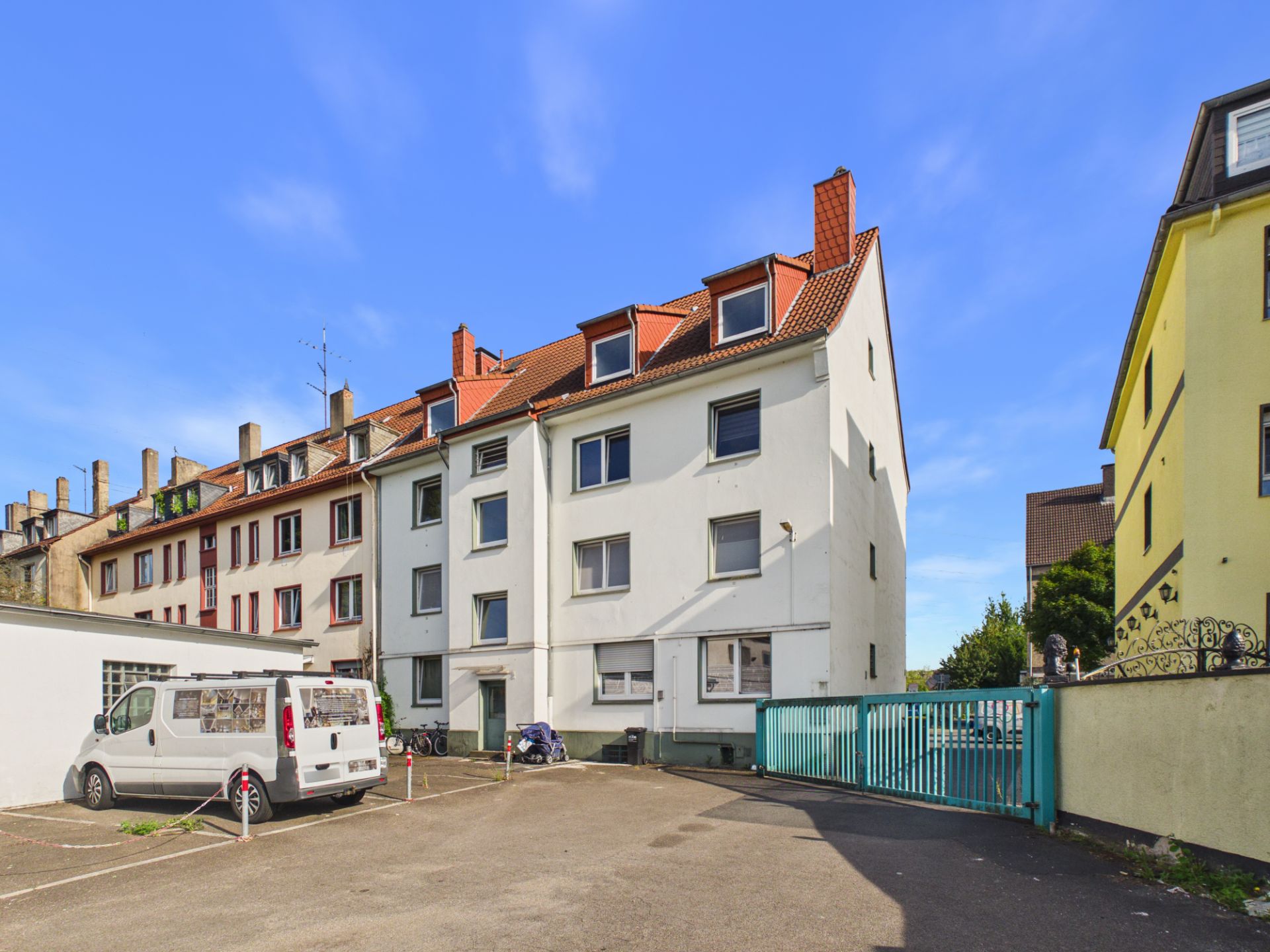 Mehrfamilienhaus mit rotem Dach und Parkplatz im Vordergrund bei sonnigem Wetter.
