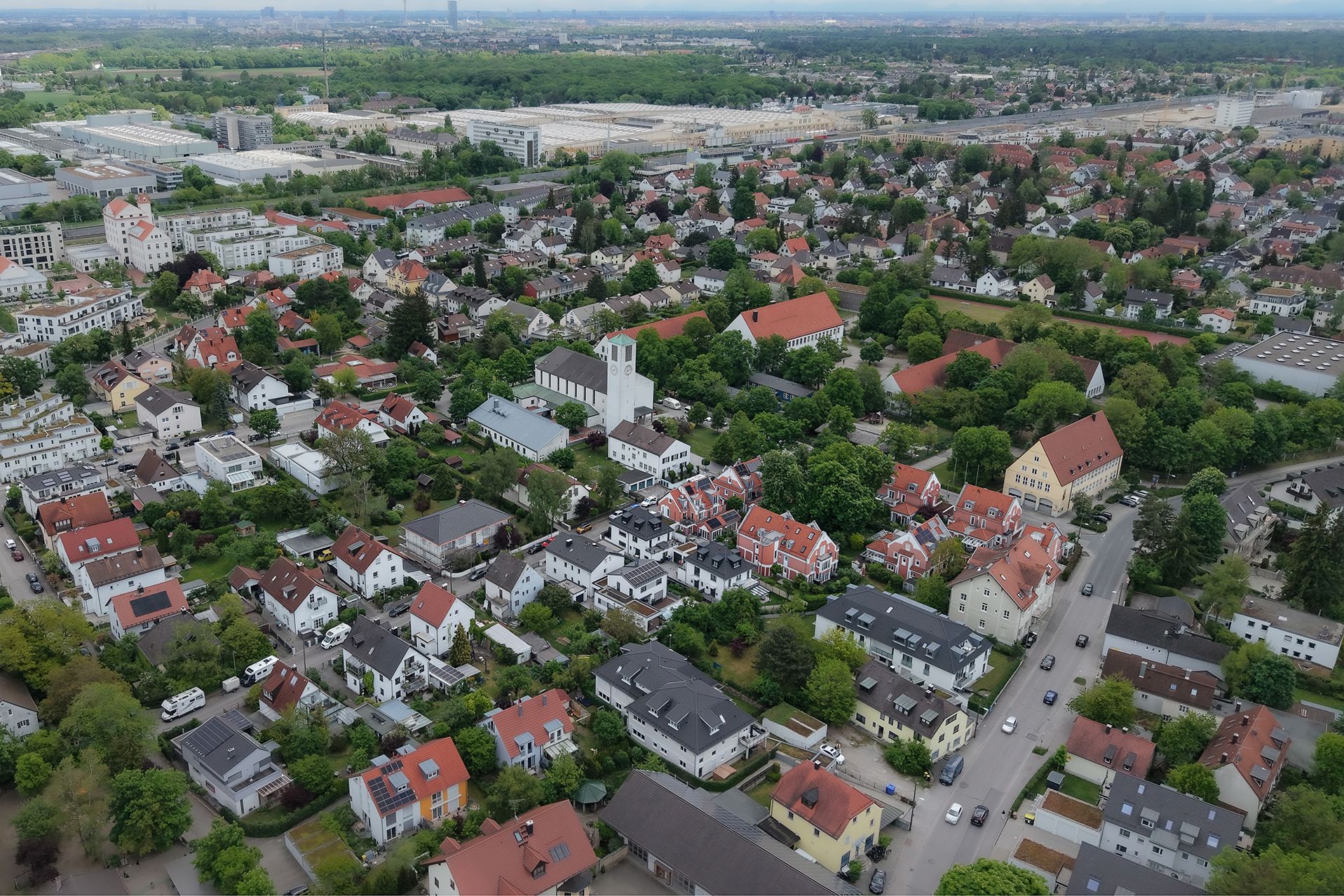 Luftaufnahme eines Wohngebiets mit vielen Häusern, viel Grün und einer Kirche in der Mitte.