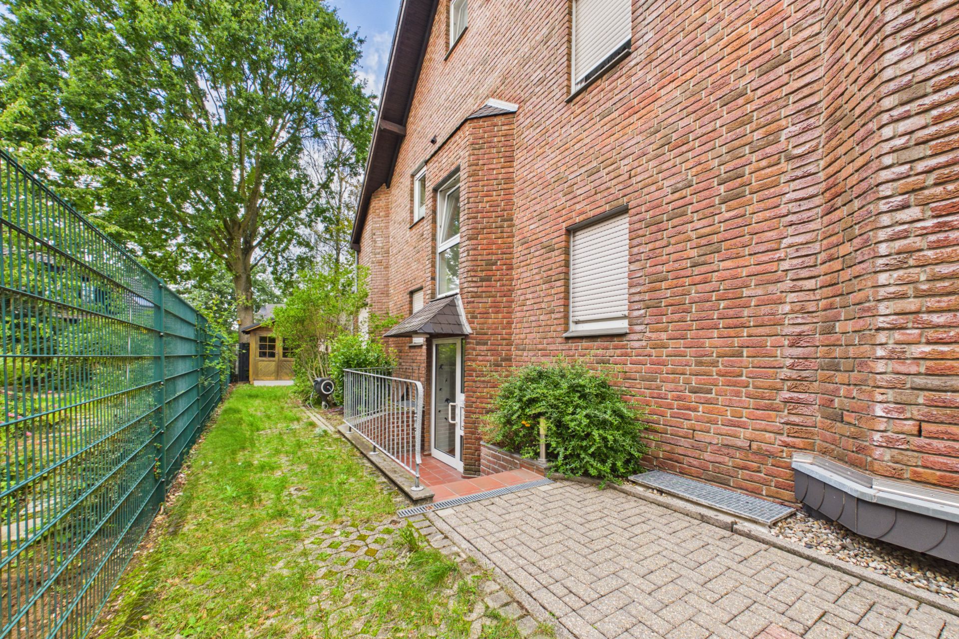 Backstein-Gebäude mit Eingangstür, Gehweg, Zaun und Bäumen im Garten.