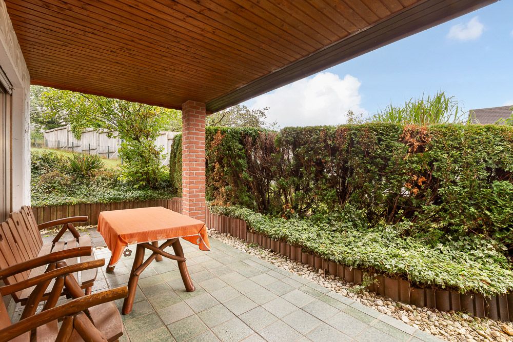 Überdachte Terrasse mit Holzbänken, Tisch mit orangener Decke und Blick auf Gartenhecke.
